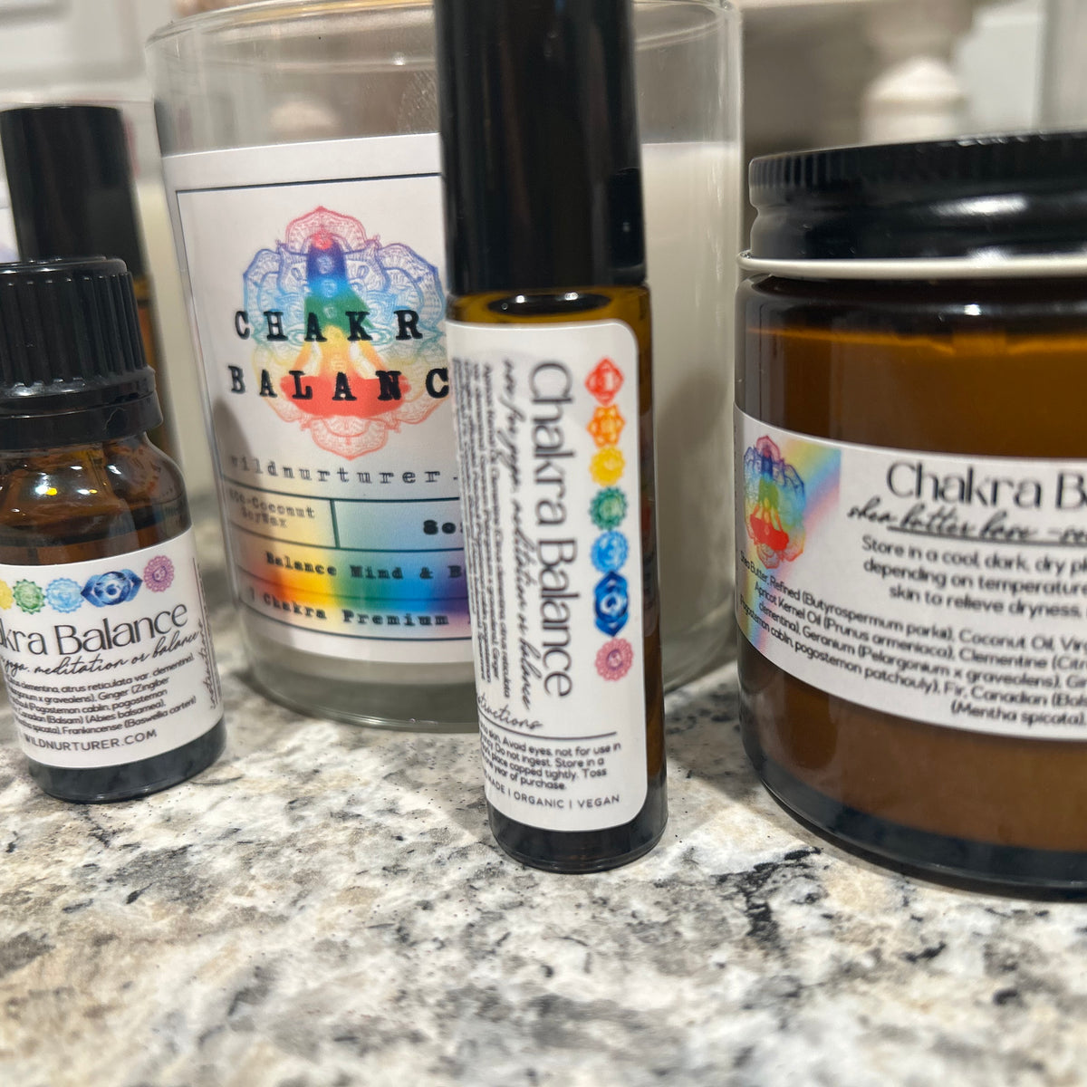 A selection of Wild Nurturer Aromatherapy’s Chakra Balance Aromatherapy - 7 Aromatic Essences (Bulk Pack of 6), including a small essential oil bottle, a roller, a jar, and a candle for aromatherapy and chakra balancing, displayed on a speckled countertop.