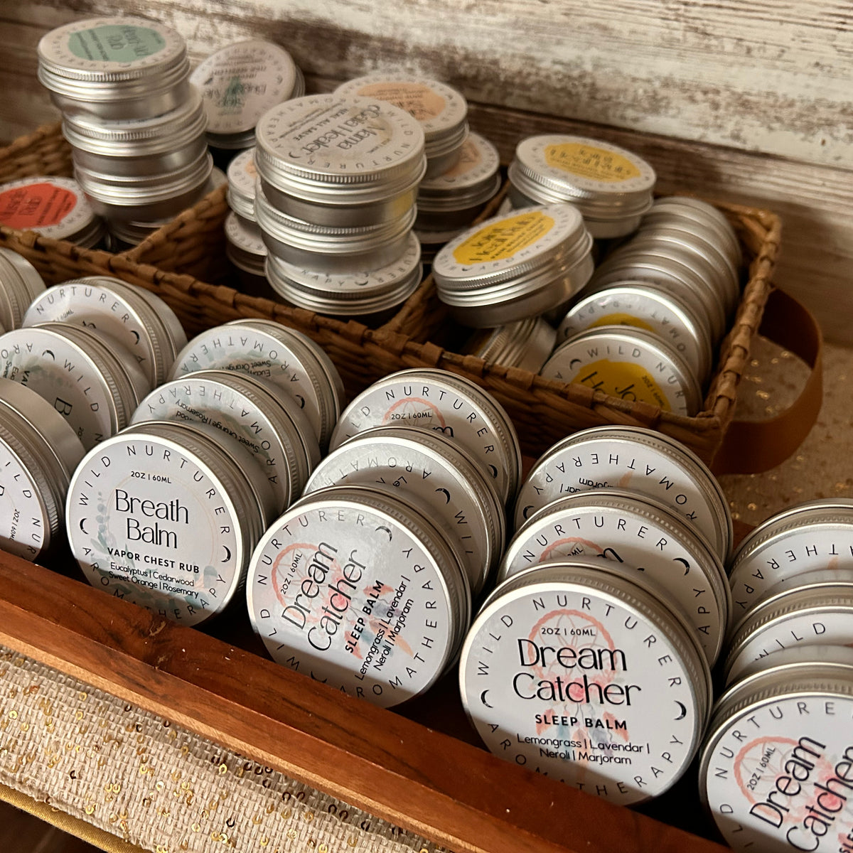 Bulk packs of “Breathe - Eucalyptus + Cedarwood Chest Balm” by Wild Nurturer Aromatherapy, featuring organic herbal formulas designed for respiratory health, are neatly arranged in rows within wooden and wicker trays on a countertop.