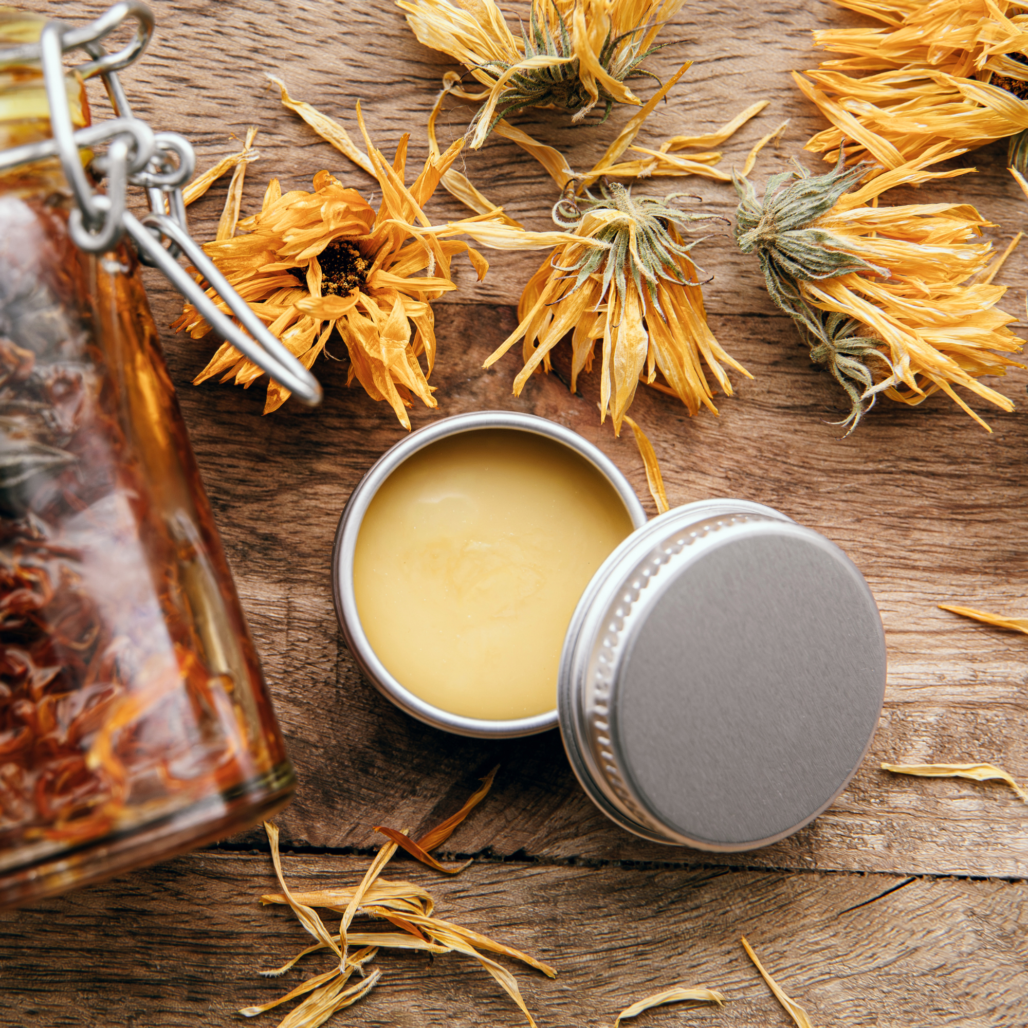 A small metal tin filled with yellow balm sits open on a wooden surface, surrounded by dried yellow flowers and a glass jar with similar contents.