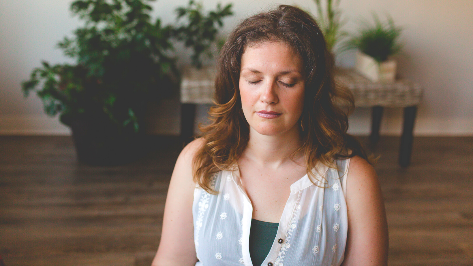 A woman with wavy brown hair sits indoors with eyes closed, wearing a sleeveless white top. There are green plants and wooden flooring in the background.