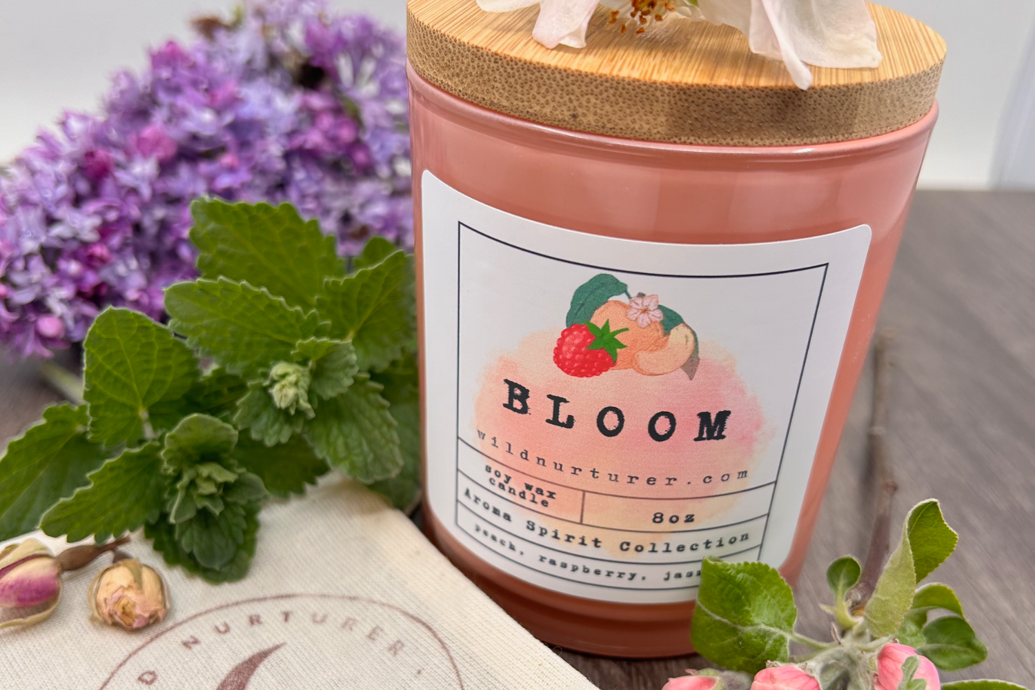 A pink scented candle labeled "Bloom" surrounded by fresh flowers and herbs on a wooden surface.