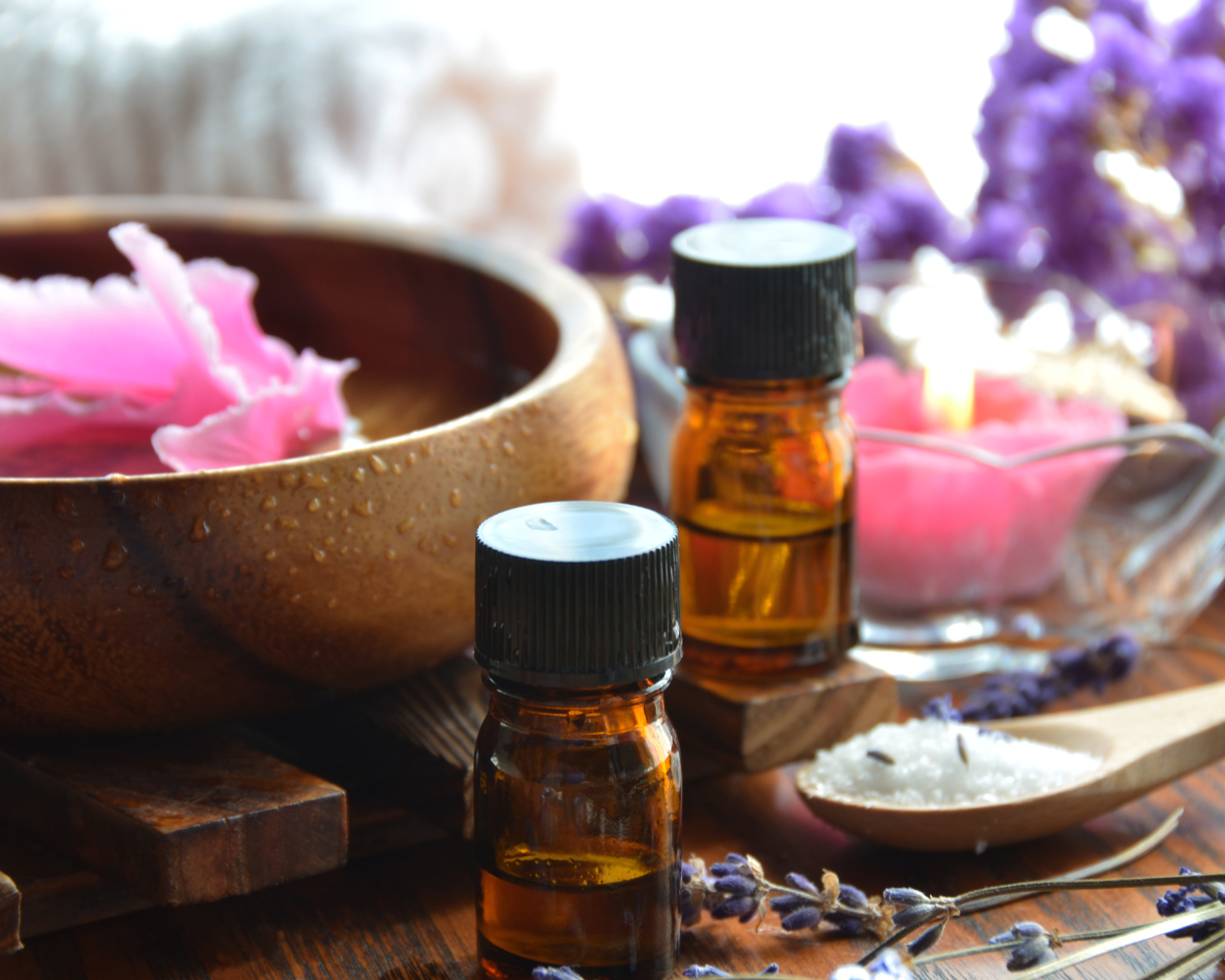 Aromatherapy setup with essential oil bottles, a bowl of flower petals, lavender, and bath salts on a wooden surface.