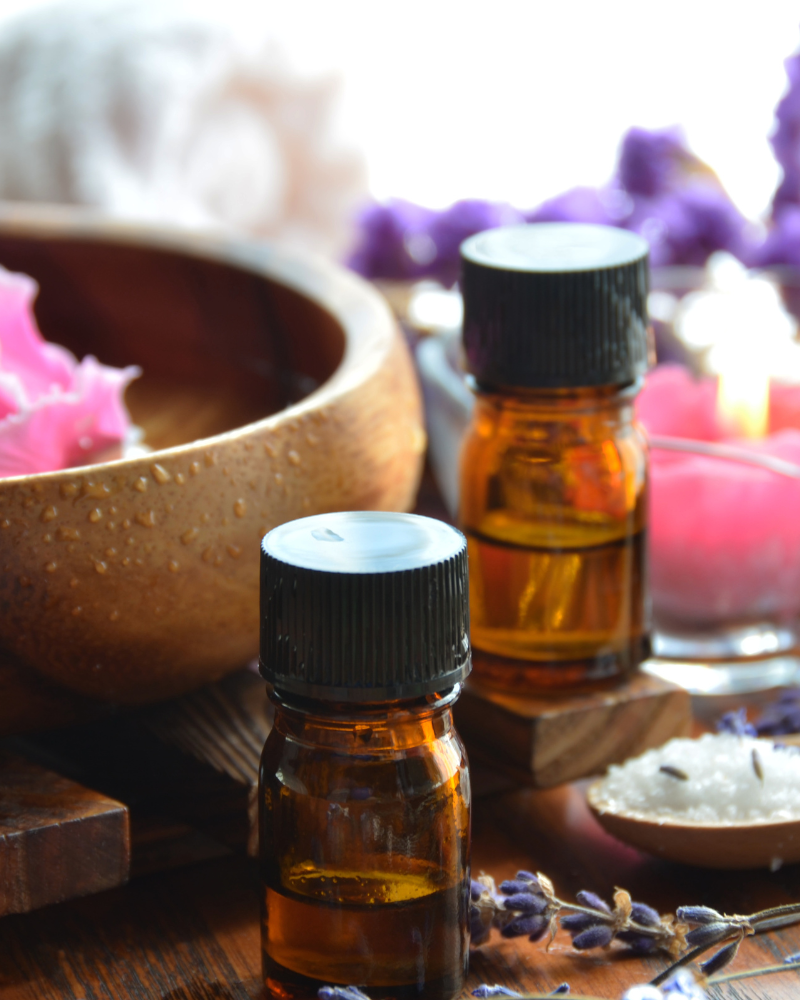 Aromatherapy setup with essential oil bottles, a bowl of flower petals, lavender, and bath salts on a wooden surface.