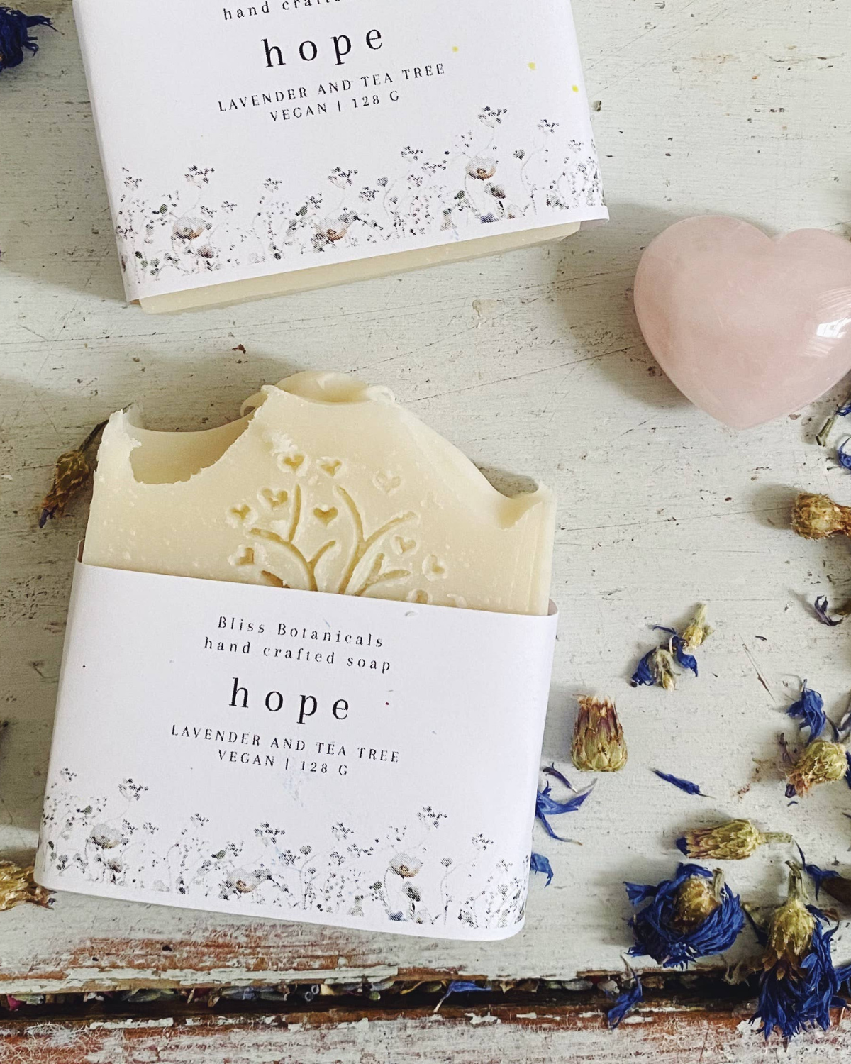 Handcrafted soap labeled "hope" with lavender and tea tree, surrounded by dried flowers and a pink heart-shaped stone on a distressed wooden surface.