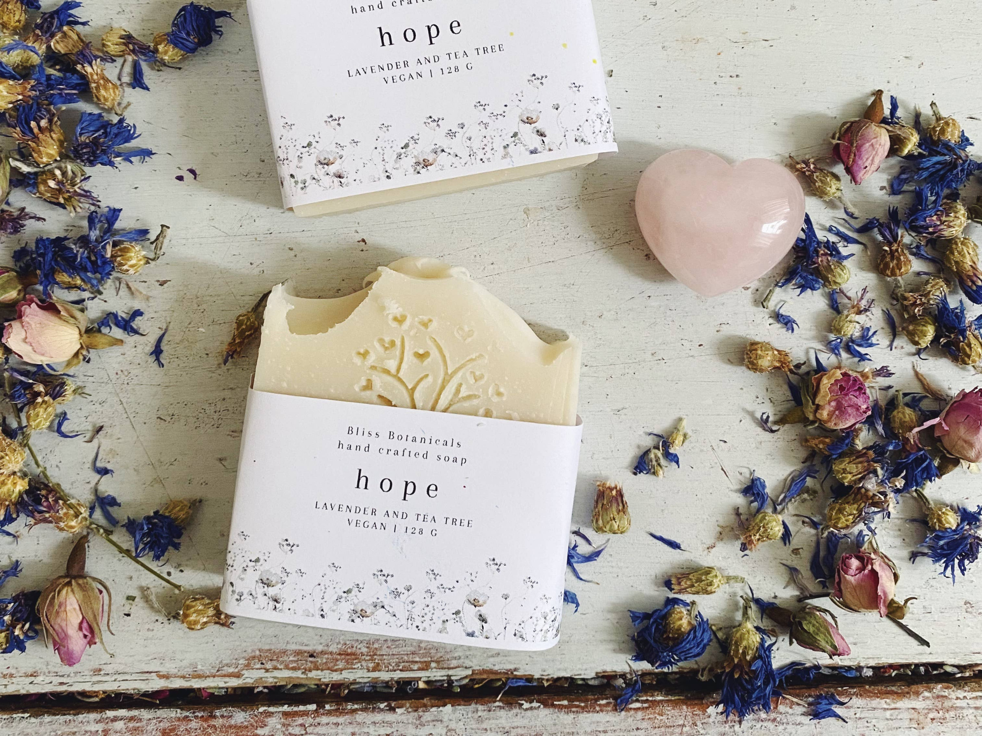 Handcrafted soap labeled "hope" with lavender and tea tree, surrounded by dried flowers and a pink heart-shaped stone on a distressed wooden surface.