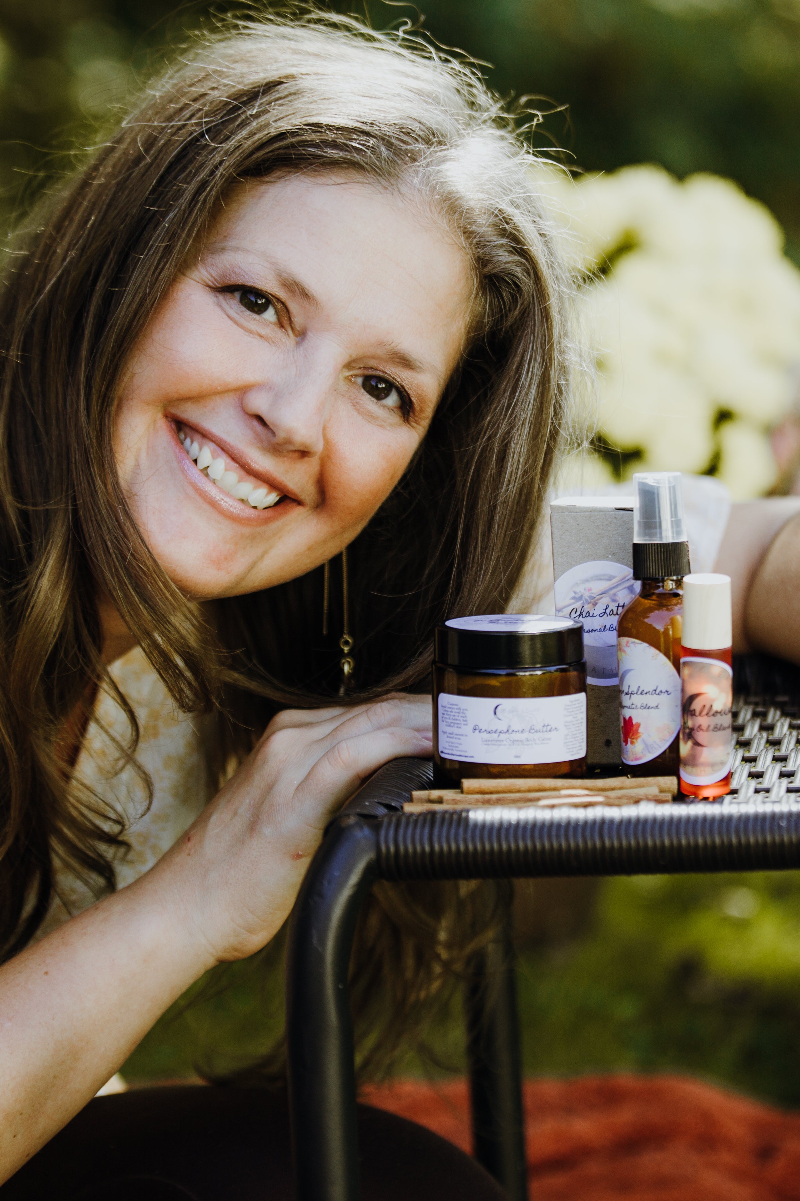 A woman with long brown hair smiles at the camera, leaning on a table displaying various skincare products in jars and bottles. The background is outdoors with greenery.