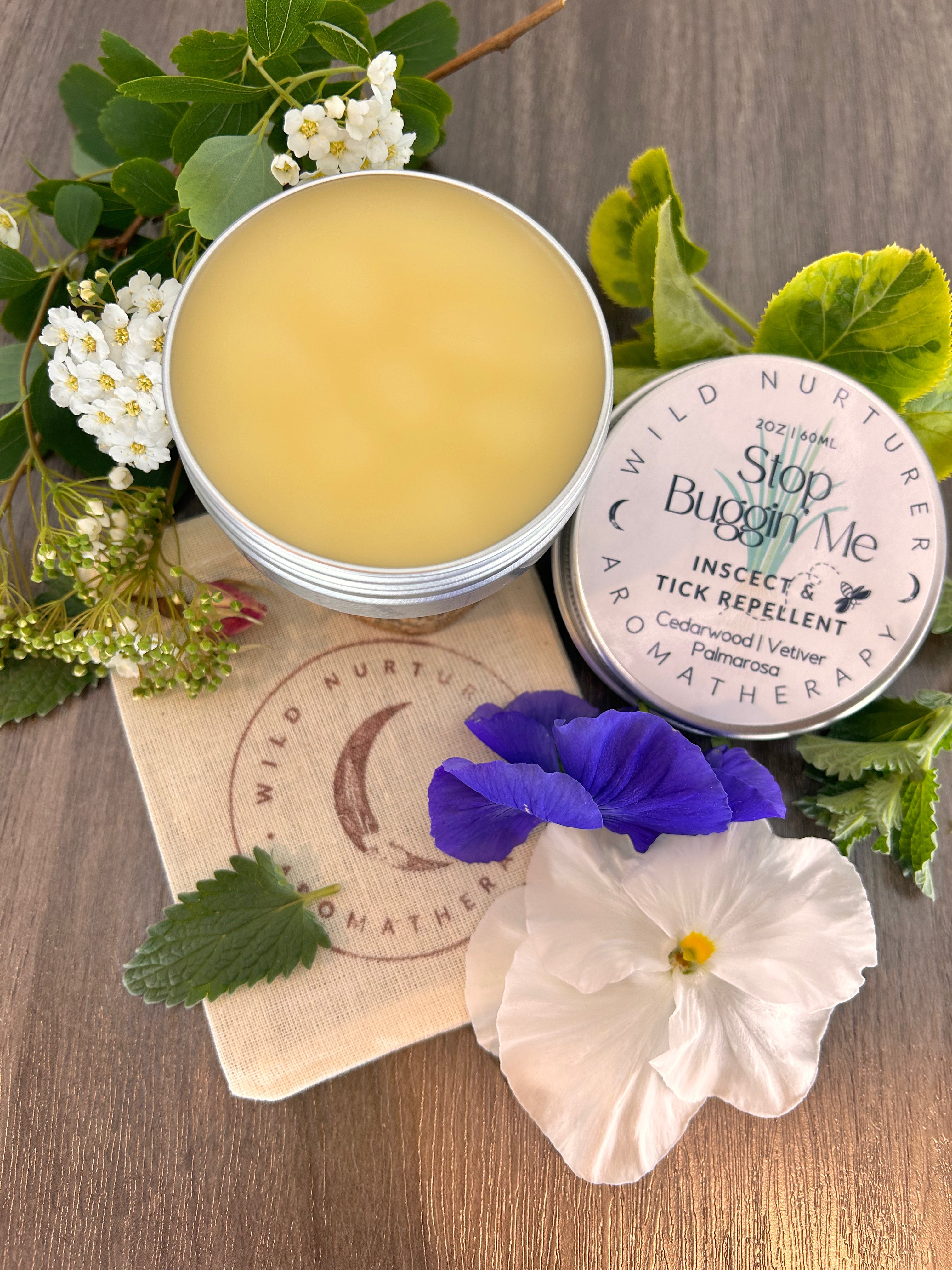 Three tins of aromatherapy salves—Joint Heal Rub, Mama Gaia Healer, and Muscle Rub—are arranged on a beige fabric, partially surrounded by green leaves.