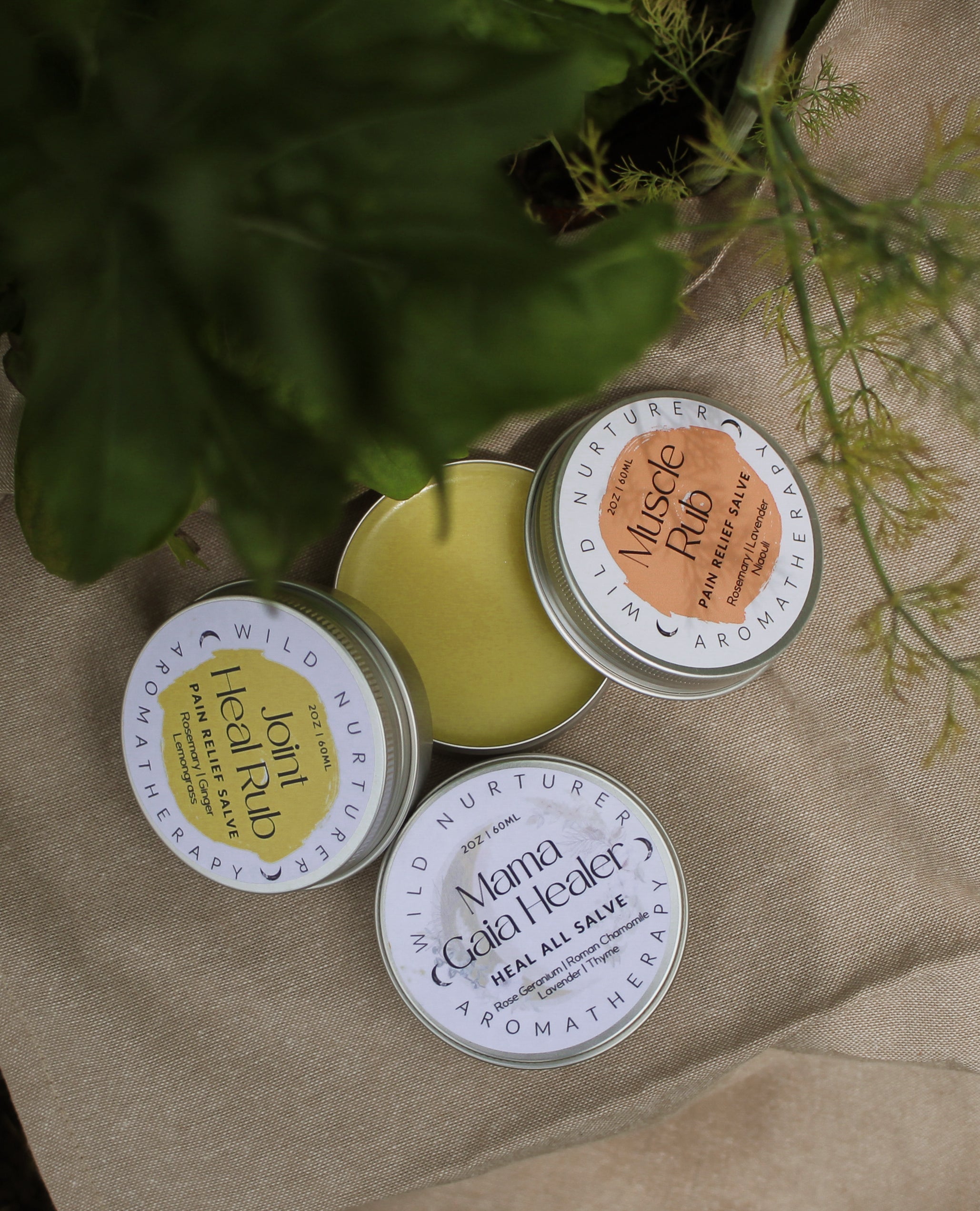Three tins of aromatherapy salves labeled “Joint Healing Rub,” “Muscle Rub,” and “Mama Gaia Healer Heal All Salve” are arranged on a beige cloth with green leaves partially visible.