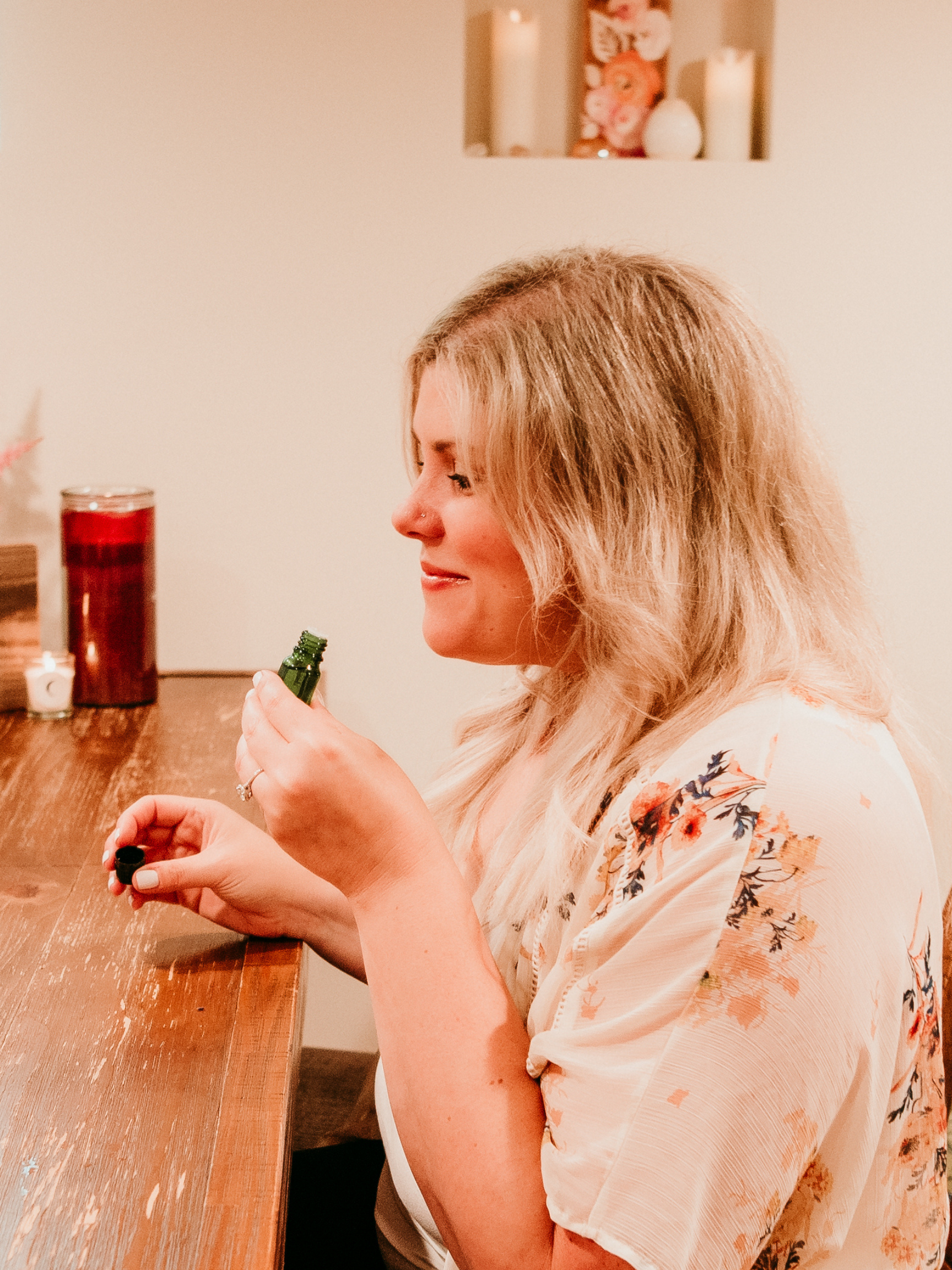 A woman with blonde hair sits at a wooden table holding a small green bottle. Lit candles and a decorative object are visible on the table and in the background.