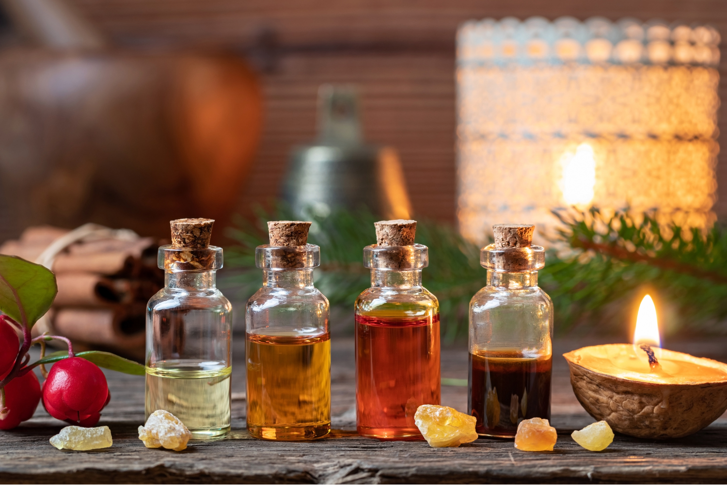 Four small glass bottles with various colored oils, a burning candle, and dried herbs on a wooden table, creating a cozy ambiance.