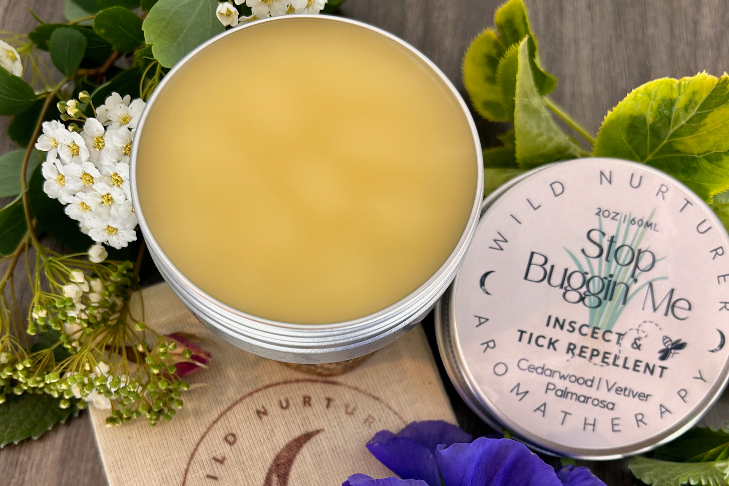Open tin of natural insect repellent balm with cedarwood and palmarosa, surrounded by fresh flowers on a wooden surface.