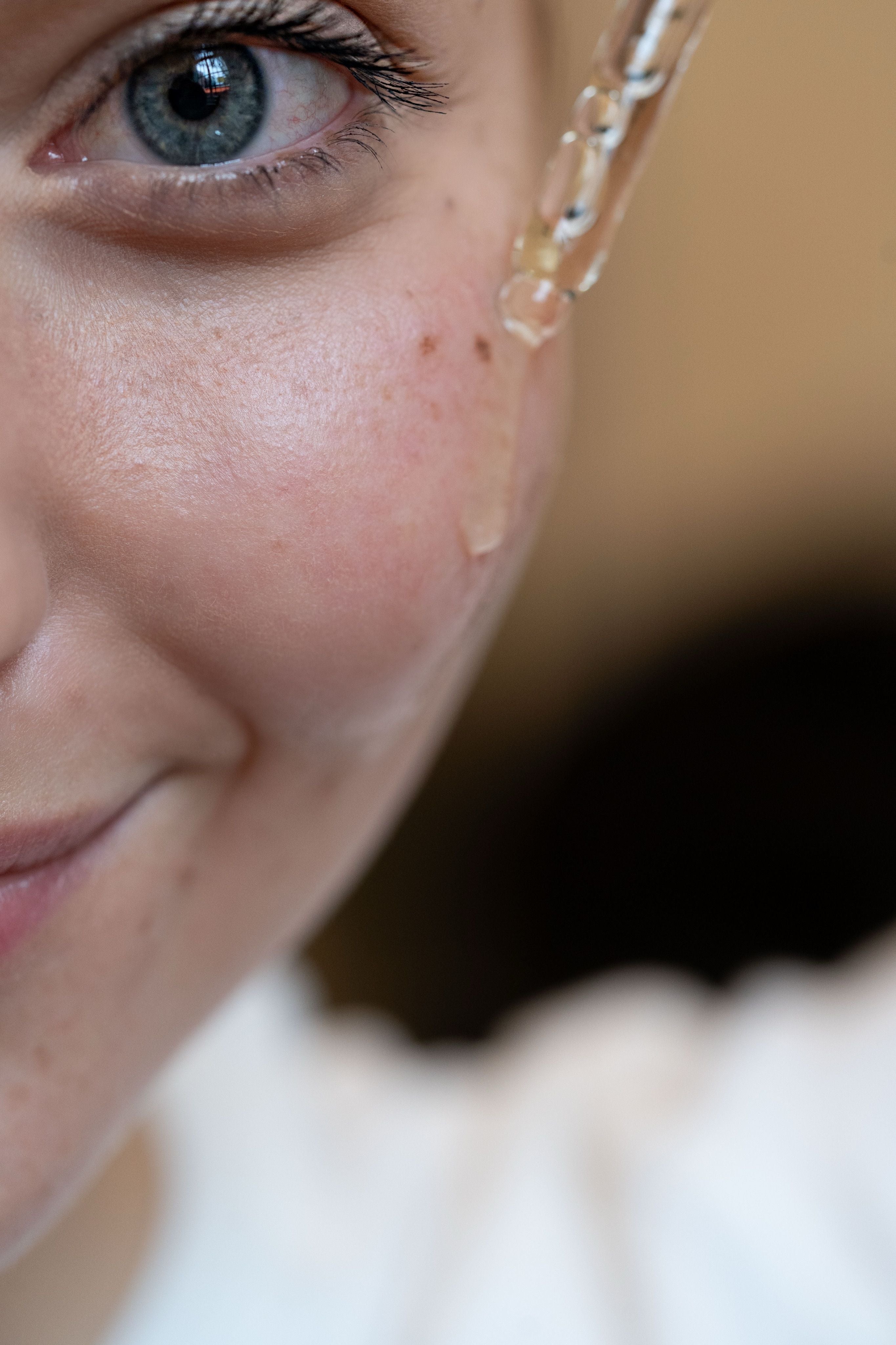 A close-up of a person’s face with a dropper applying serum or oil to their cheek. The person has blue eyes and light skin. Only part of the face is visible.