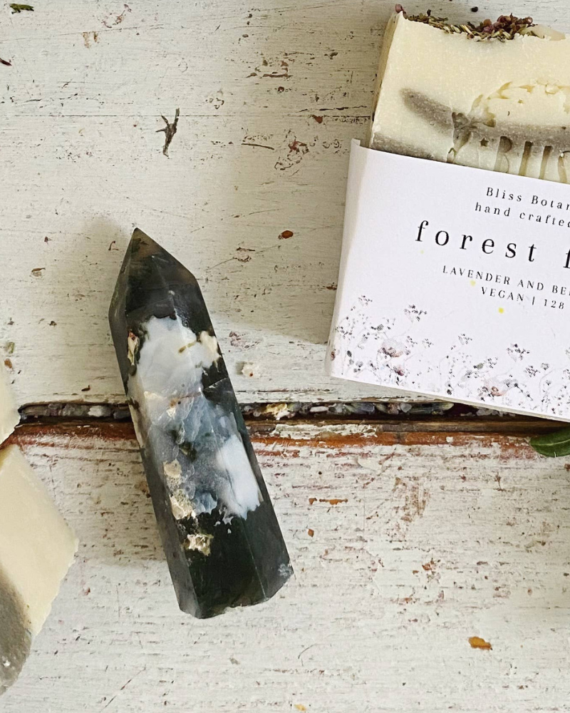 Handmade soap bars with dried herbs on a white wooden surface next to a "forest fairy" soap packaging and a green plant leaf.