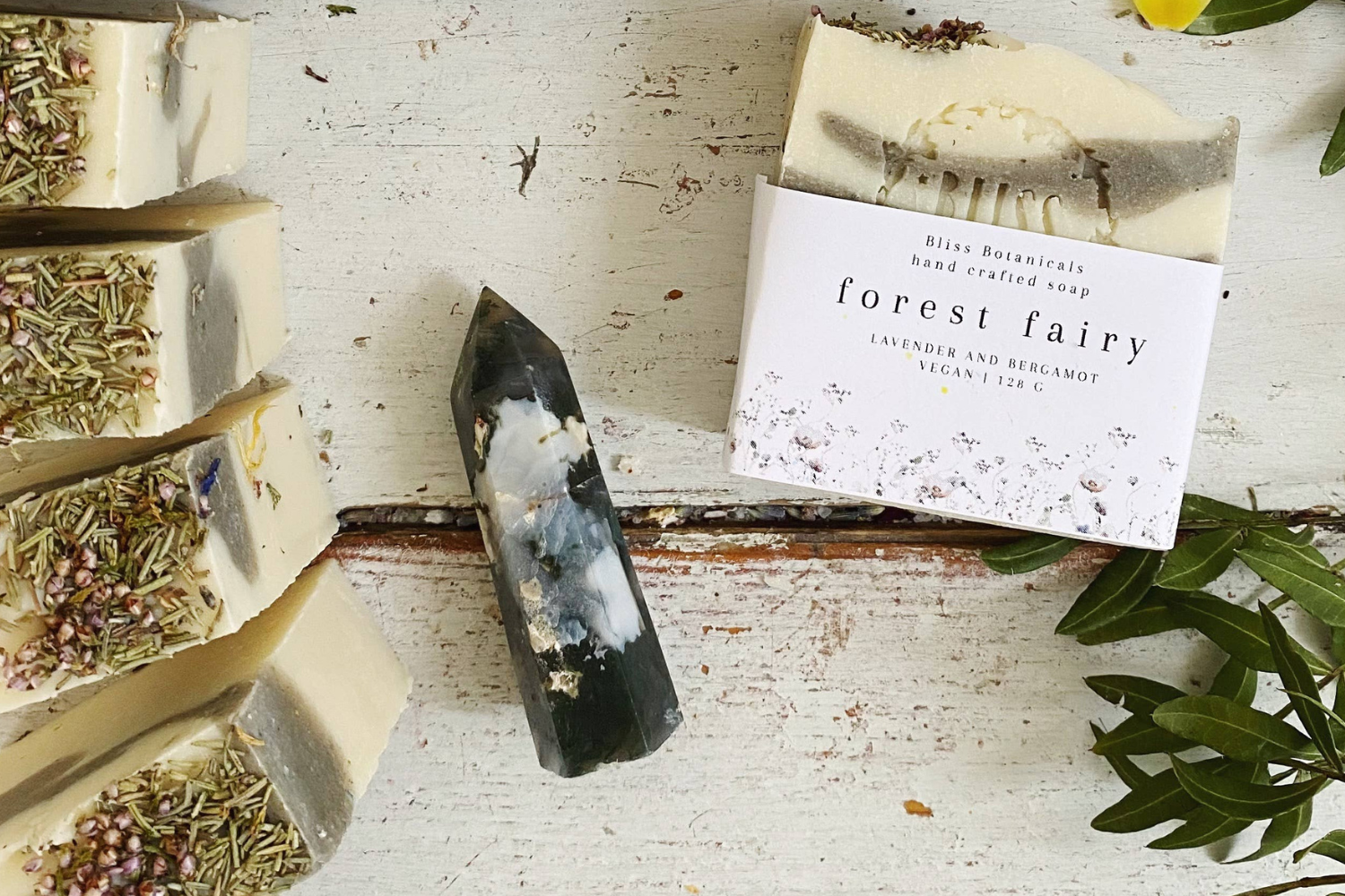 Handmade soap bars with dried herbs on a white wooden surface next to a "forest fairy" soap packaging and a green plant leaf.