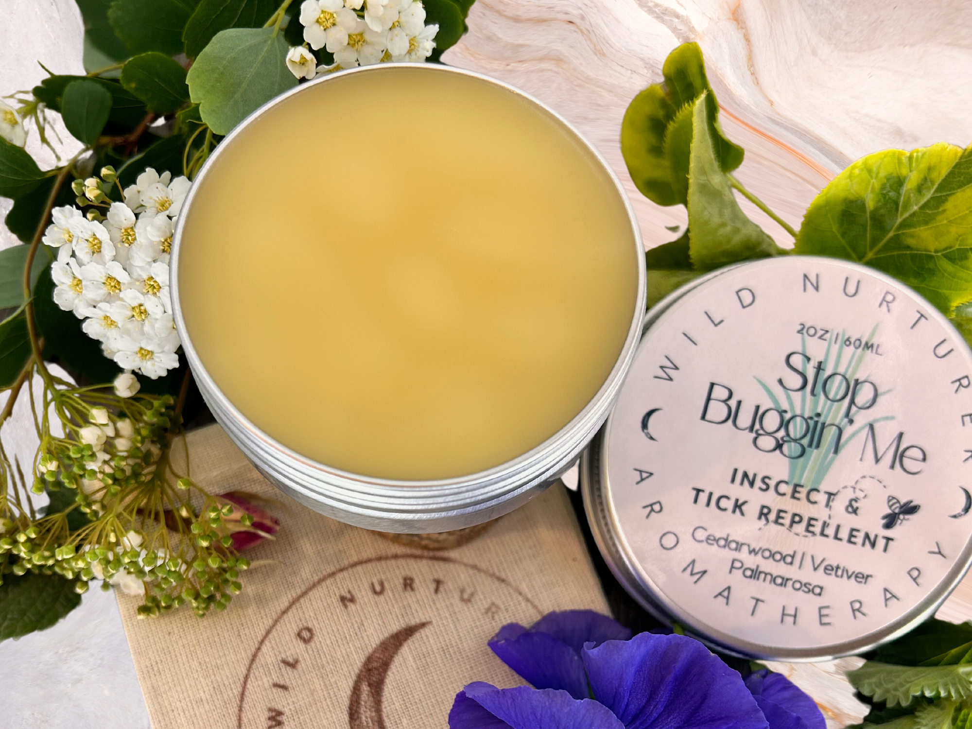 Open jar of natural tick repellent cream surrounded by flowers and leaves on a tabletop. .
