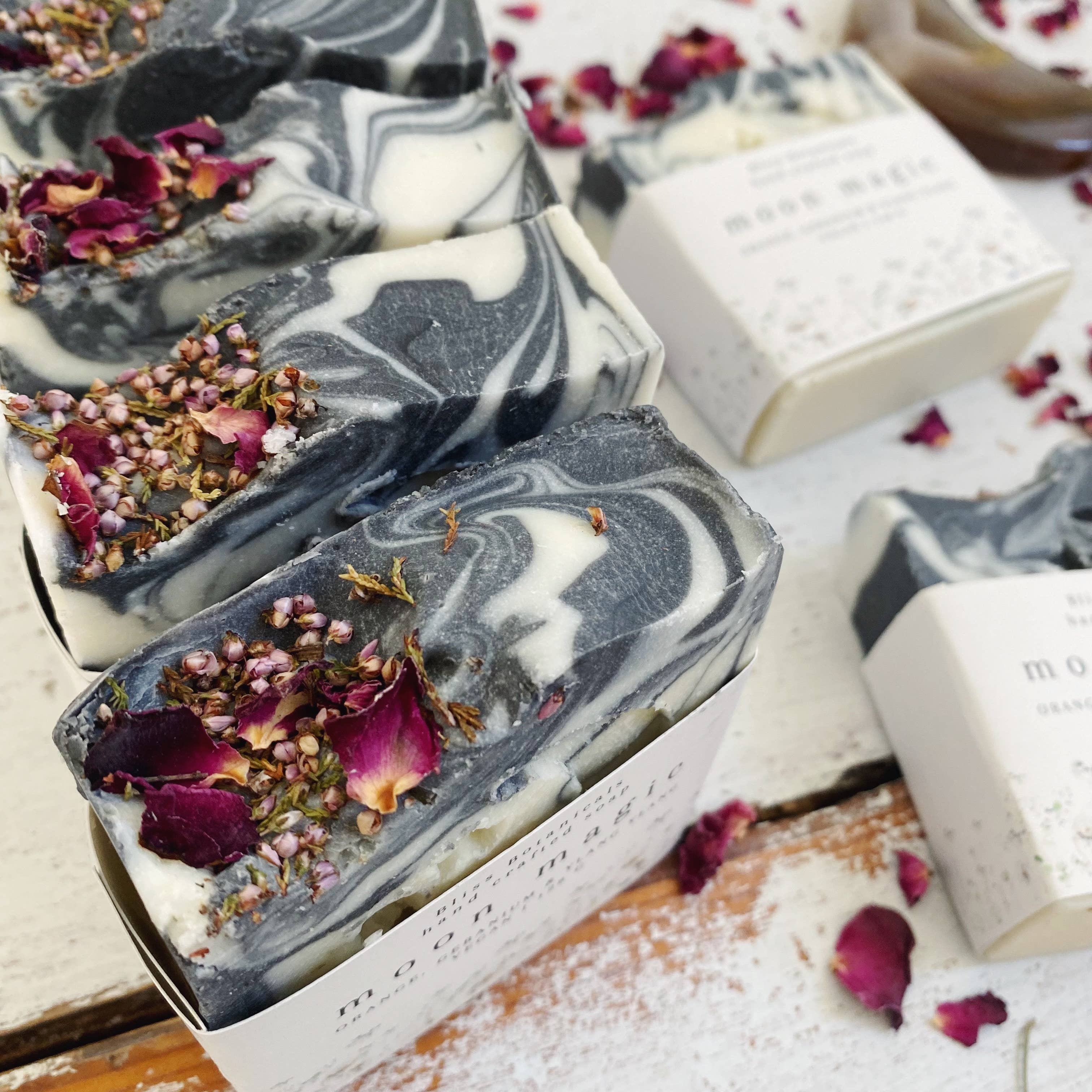 Moon Magic Soap bars with marbled patterns alongside dried rose petals, crystals, and a crescent moon dish on a rustic white wooden surface, infused with wild nurturer aromatherapy essential oils. Created by Bliss Botanicals.