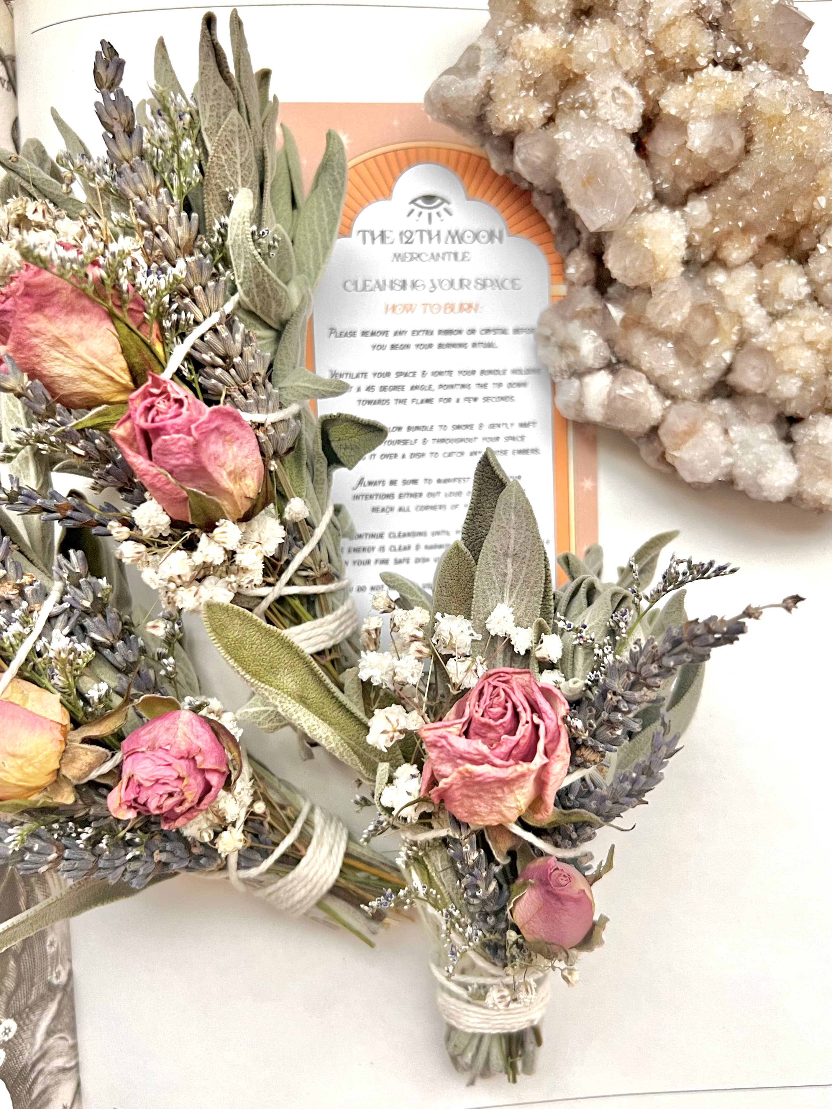 Two small floral bouquets with dried roses and lavender next to a MAGIC RITUALS Mini Sage Smudge Bundle - Smoke Cleanser on a pale background, infused with essential oil for aromatherapy benefits by The 12th Moon.