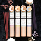 Four packages of cylindrical spell incense labeled "Self Love," "Prosperity," "Happiness," and "Friendship" are surrounded by incense sticks and dried flowers. Each package, from the Something Different Wholesale Set of 12 Herbal Spell Incense Stick Gift Sets in Display, features a moon-designed round ornament, making them perfect for enhancing your magickal practices.