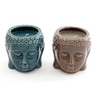 Two 10cm Buddha Head Candle Pots from Something Different Wholesale, featuring ceramic holders shaped like Buddha heads—one in green and the other in brown—each with unlit scented candles inside, offering a touch of tranquility and home fragrance.