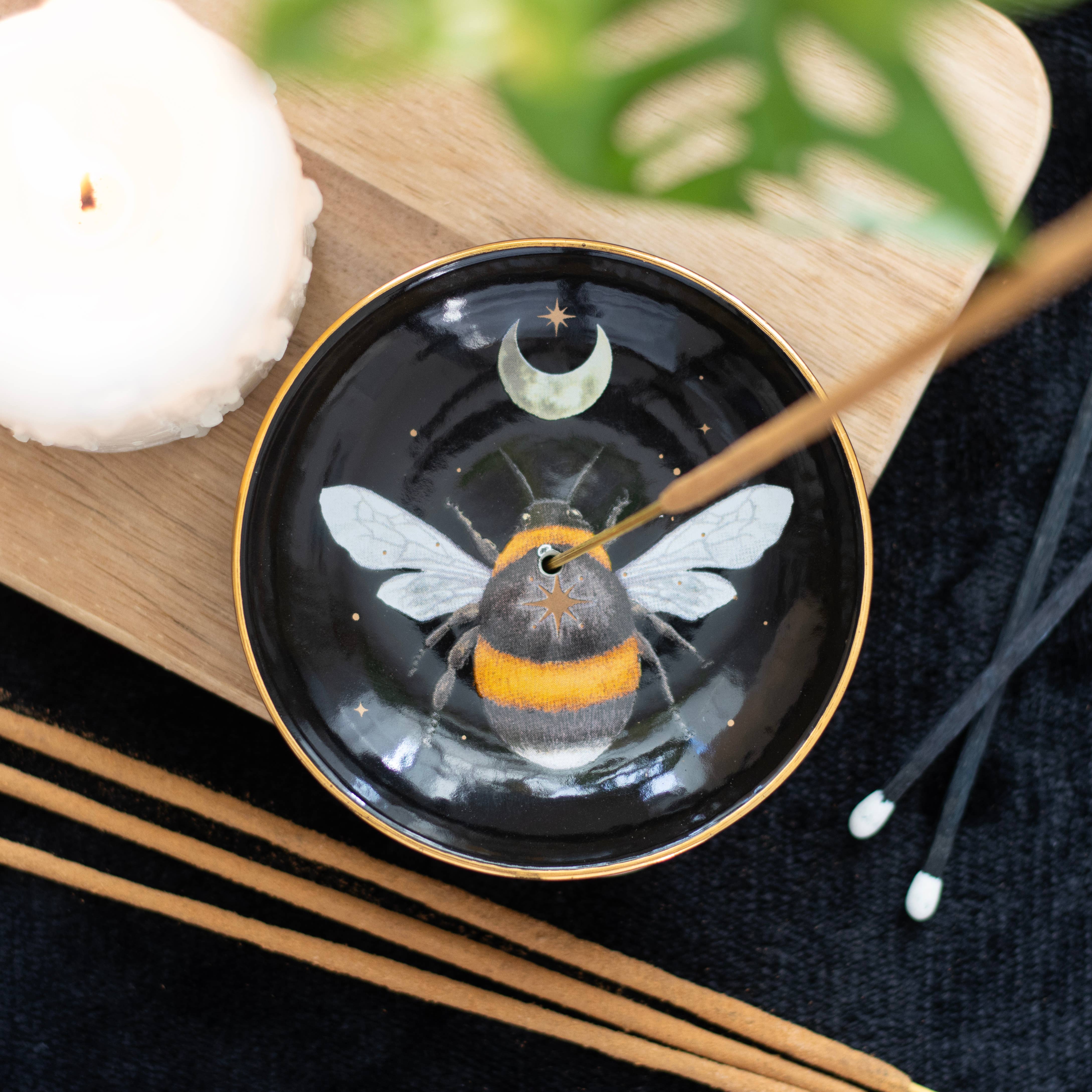 The Forest Bee Ceramic Incense Holder Plate by Something Different Wholesale features a painted forest bee and crescent moon design. An incense stick is inserted into the dish, which rests on a wooden surface alongside a lit candle and several other incense sticks—ideal for those who appreciate mystical woodland gifts.