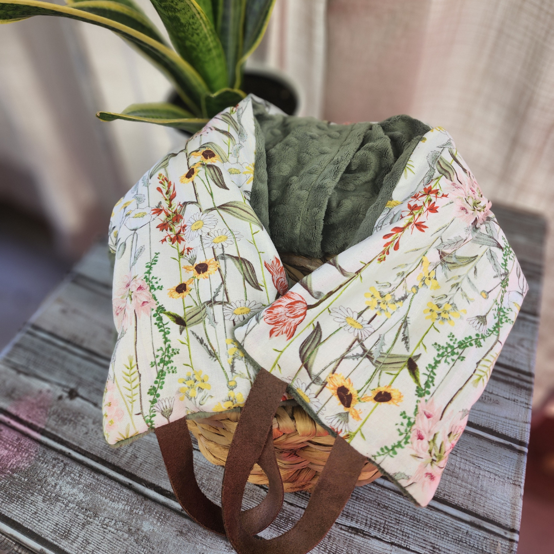 A floral-patterned scarf draped over a wicker basket on a wooden table, with a green plant in the background, evokes a serene setting where one might find the Calm Your Vibes Hot & Cold Weighted Neck Wraps with Washable Boho Wildflowers Covers, filled with a soothing rice and flaxseed mix and scented with lavender, to unwind.