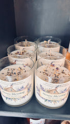 A set of six House Blessing Soy Candles by Wild Nurturer Aromatherapy, labeled “Lavender + Sage” and topped with herbs, are arranged on a black shelf. Crafted as aromatherapy candles, this bulk pack features soothing scents of lavender and sage to promote peace and positive energy in any space.