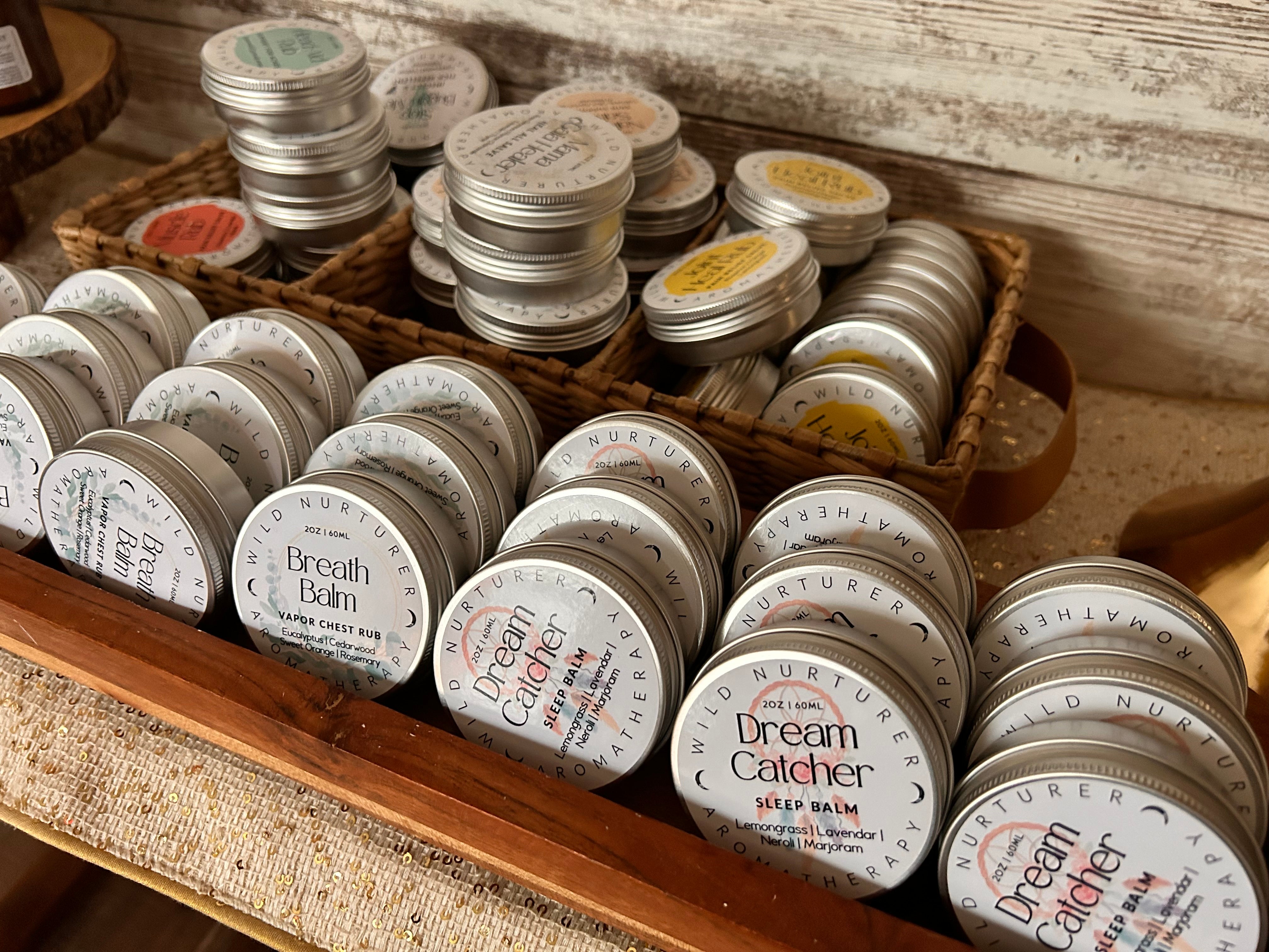 Bulk packs of “Breathe - Eucalyptus + Cedarwood Chest Balm” by Wild Nurturer Aromatherapy, featuring organic herbal formulas designed for respiratory health, are neatly arranged in rows within wooden and wicker trays on a countertop.
