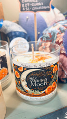 A glass candle from the Moon Medicine Moon Phase Candle Collection by Heaven & Earth Aromatherapy is labeled “Waning Moon,” featuring an illustrated moon and orange flowers on the label, and is shown among other decorative items.