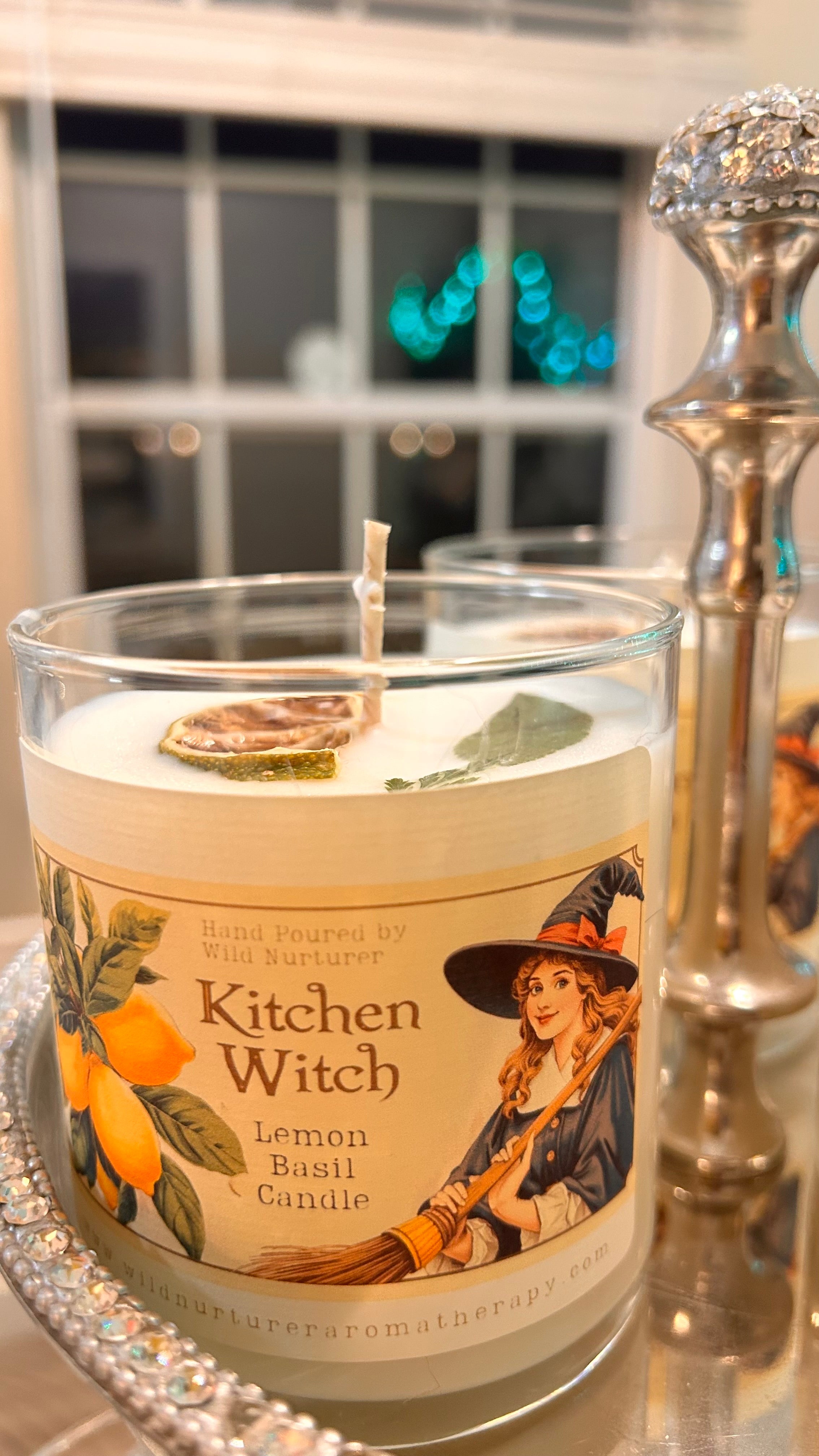 A Wild Nurturer Aromatherapy “Kitchen Witch Soy Candle – Lemon + Basil” is displayed on a tray, topped with a dried lemon slice and basil leaf. This odor neutralizing lemon basil candle sits beside a crystal candle holder as twinkling lights shine through the window in the background.