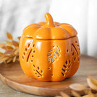 An Orange Autumn Leaves Fall Pumpkin Oil Burner by Something Different Wholesale, featuring intricate cut-out leaf patterns, is set on a wooden surface with dried leaves in the background, perfect for adding an autumnal touch to your decor.