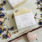 Two Hope - Lavender and Tea Tree Cold Pressed Soap bars with floral embossing, accompanied by dried flowers and a pink heart-shaped stone, displayed on a worn wooden surface.