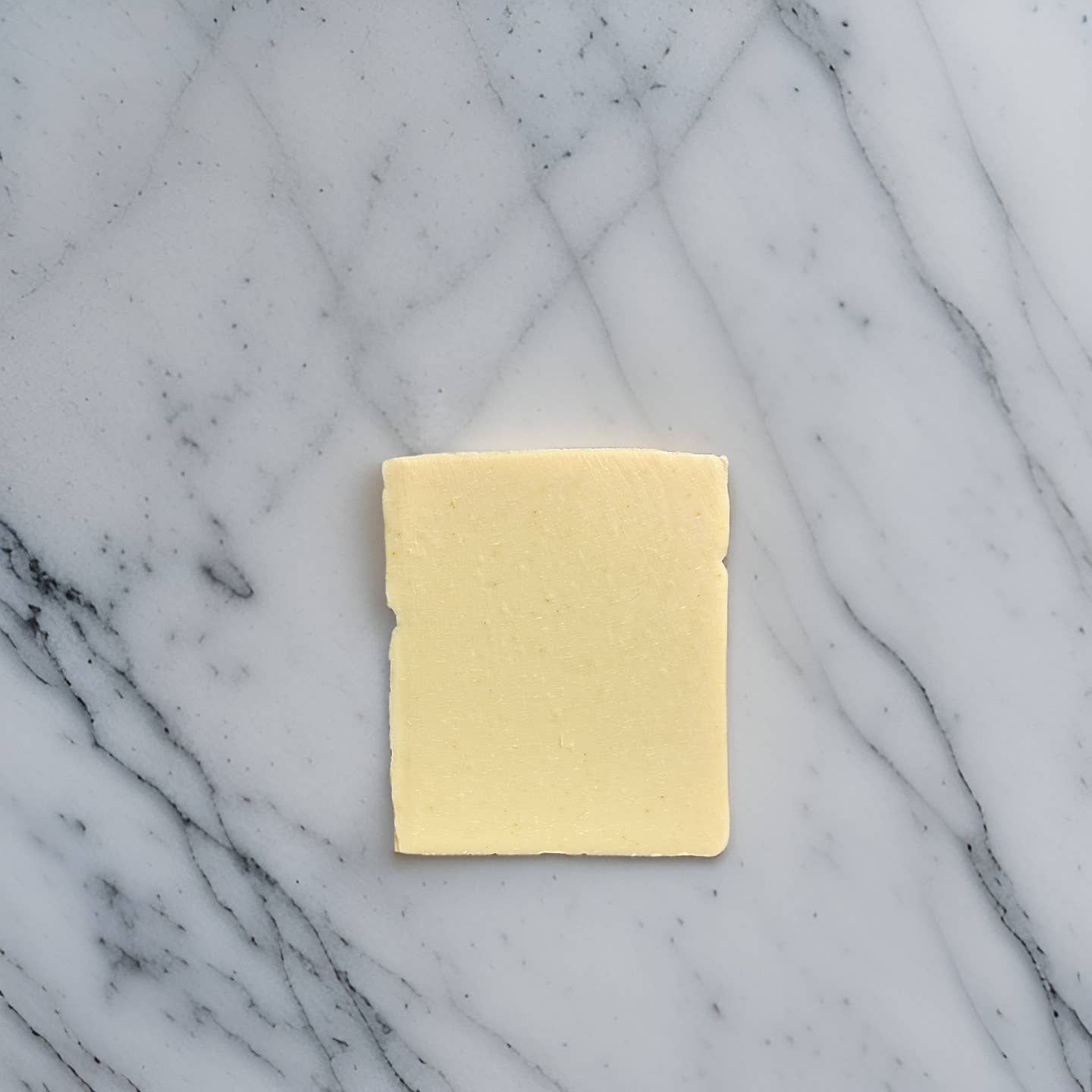 A single slice of cheese sits on a white marble surface with grey veining patterns, reminiscent of Standing Spruce's Lemongrass and Citronella Soap crafted with care.