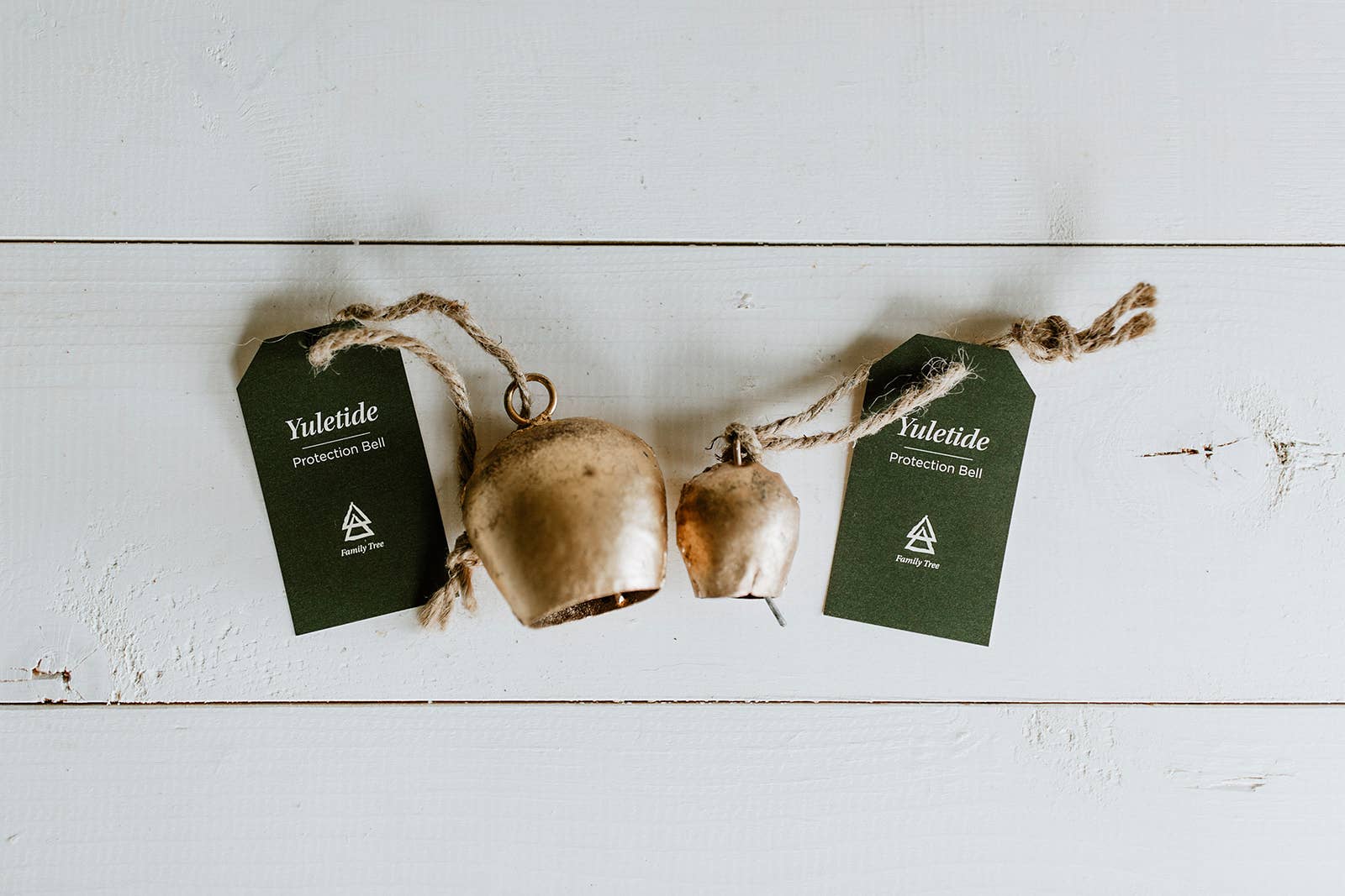 Two large rustic golden bells from Standing Spruce's "Protection Bell" collection hang by twine with green tags attached on a white wood background, warding off evil spirits.