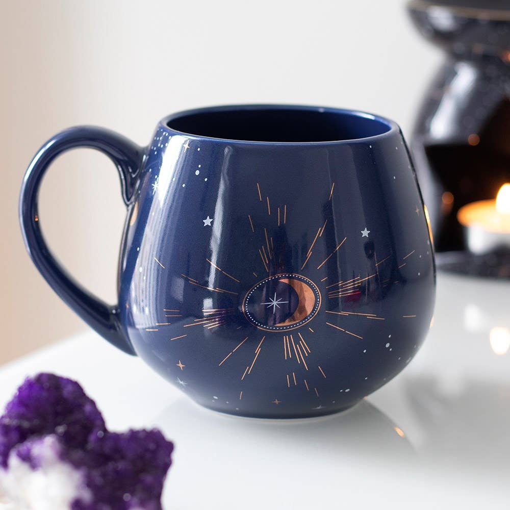 A Blue Crescent Moon Rounded Mug by Something Different Wholesale, featuring a celestial design with stars and a crescent moon illustration accented with gold foil details, sits on a white surface. An out-of-focus candle and purple crystal are in the background.