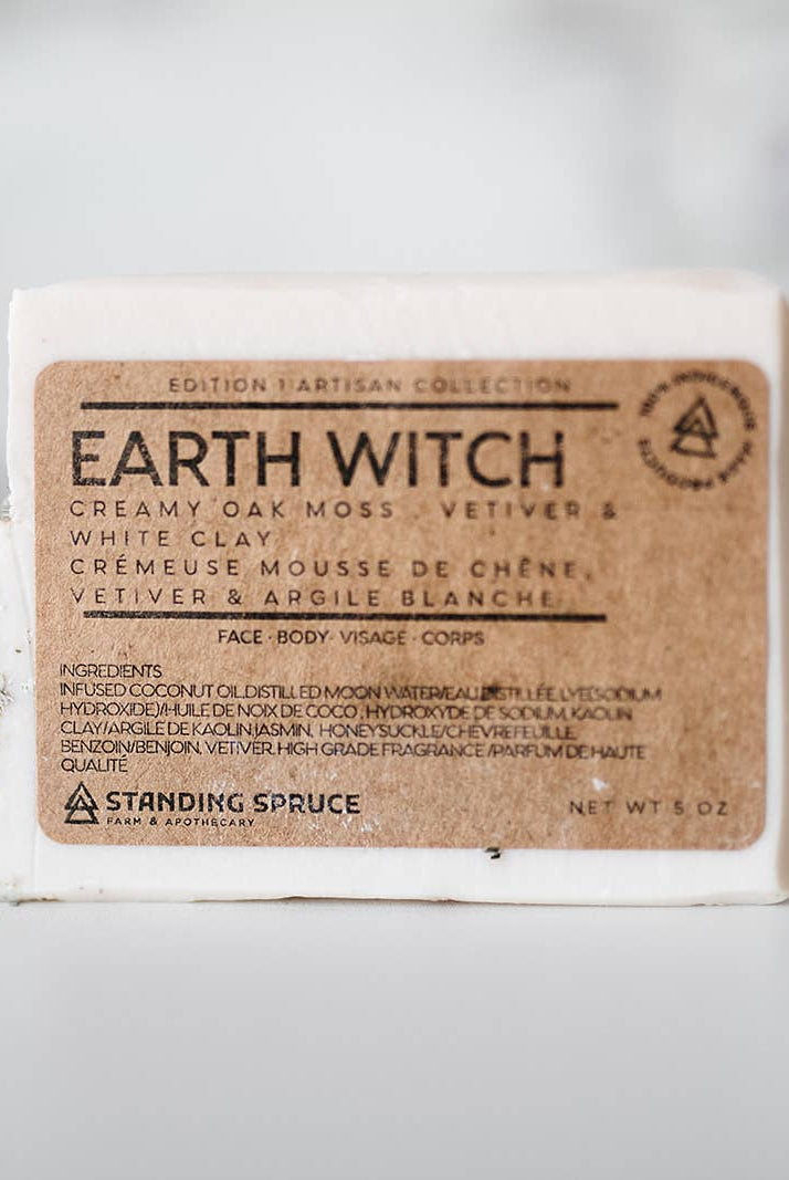 A bar of Standing Spruce "Earth Witch Soap" aromatherapy soap with text detailing ingredients and branding, placed on a blurred light background.
