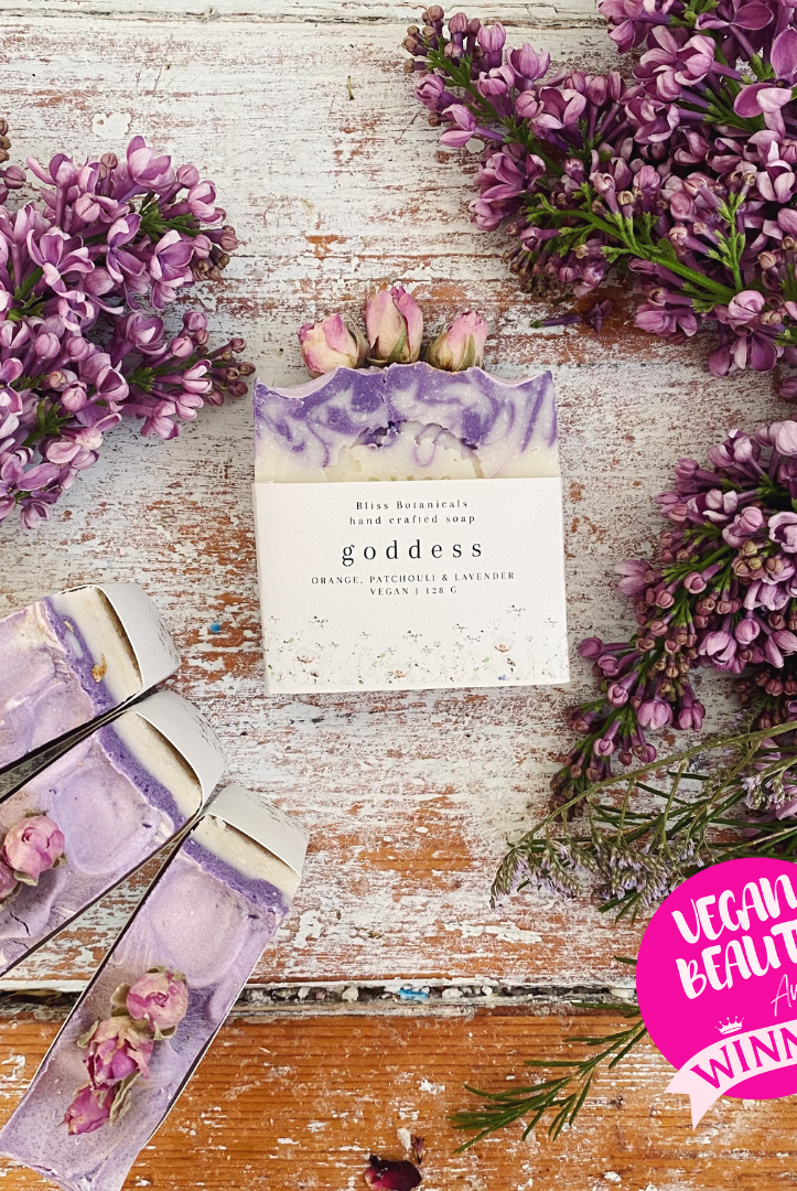 Handmade floral soap bars infused with essential oil, featuring purple lilac blooms spread across a rustic white wooden surface and adorned with a Bliss Botanicals Award Winning Goddess Soap - Orange, Patchouli & Lavender sticker.