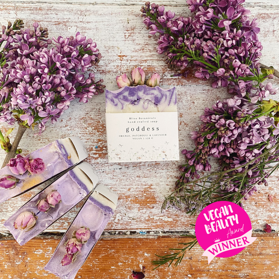 Handmade floral soap bars infused with essential oil, featuring purple lilac blooms spread across a rustic white wooden surface and adorned with a Bliss Botanicals Award Winning Goddess Soap - Orange, Patchouli & Lavender sticker.