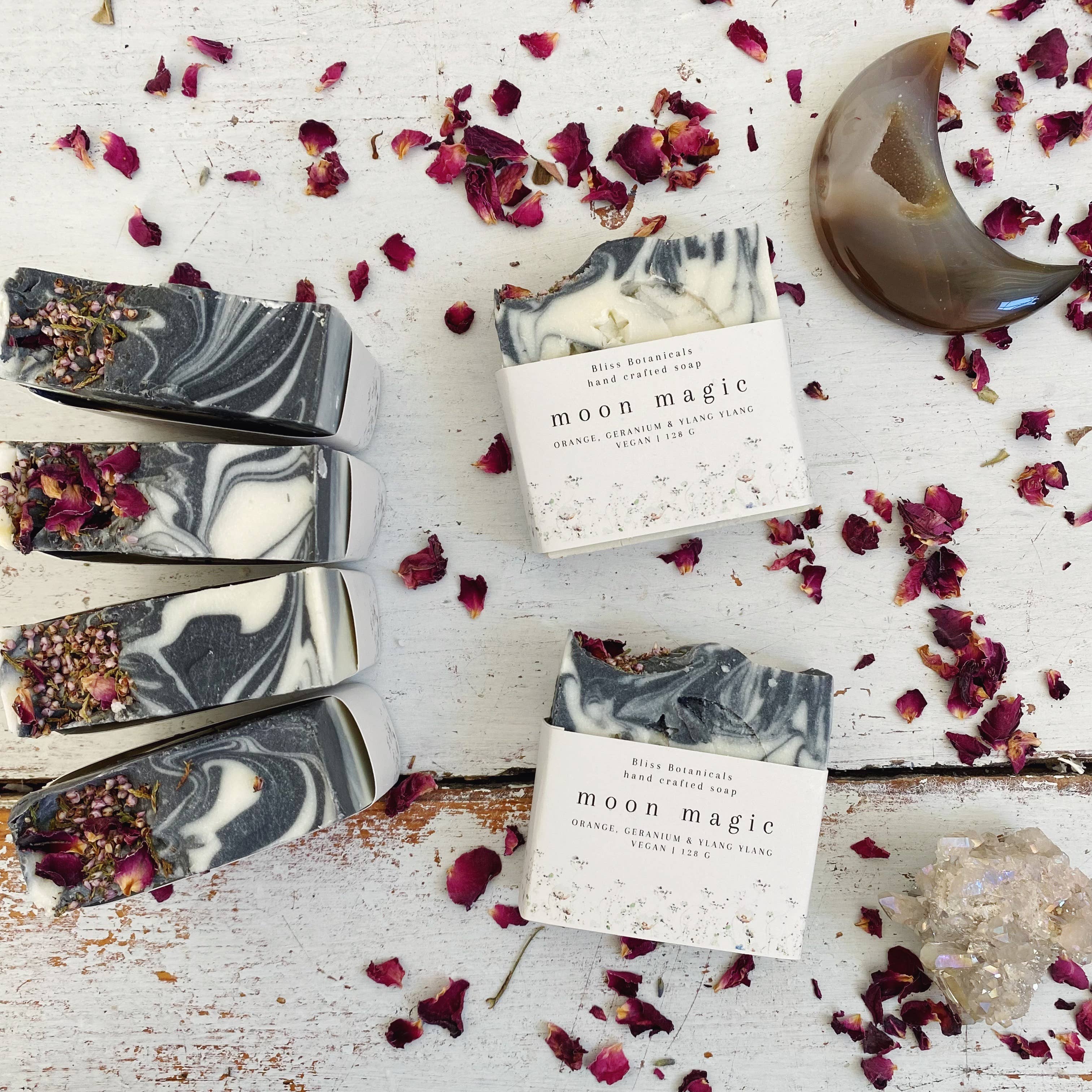 Moon Magic Soap bars with marbled patterns alongside dried rose petals, crystals, and a crescent moon dish on a rustic white wooden surface, infused with wild nurturer aromatherapy essential oils. Created by Bliss Botanicals.