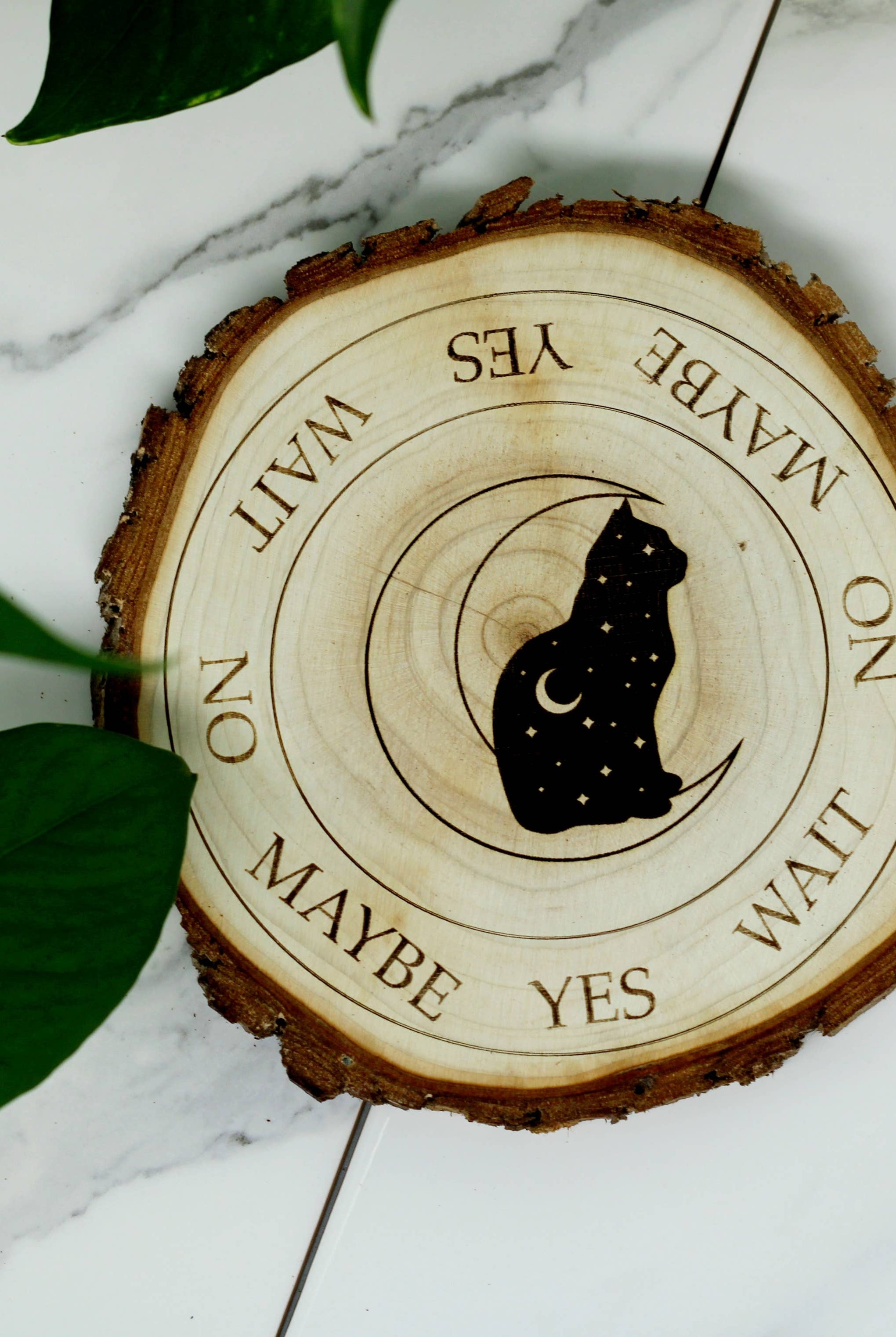 A Faire Three Witches Tea Shop Handmade Natural Wood Cat + Crescent Moon Pendulum Board & Pendulum with "maybe, yes, wait" etched around a spiraled design, infused with essential oil, placed on a marble surface beside a green leaf.