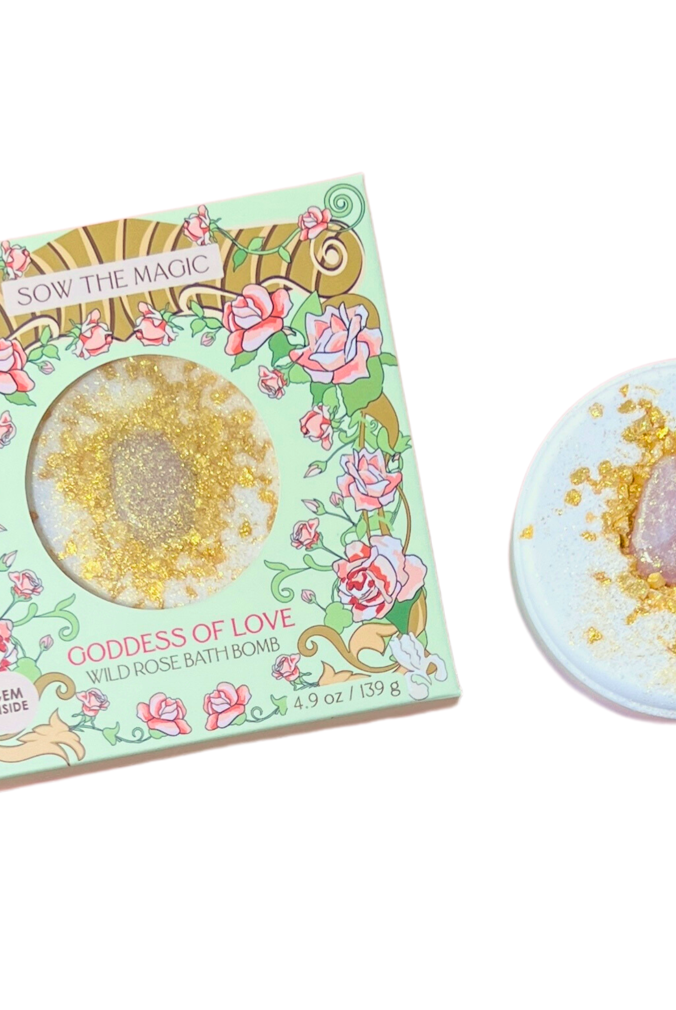 A Sow the Magic Goddess of Love Wild Rose Bath Bomb with Rose Quartz bath bomb with sparkly gold glitter and essential oil, next to its floral packaging on a white background.