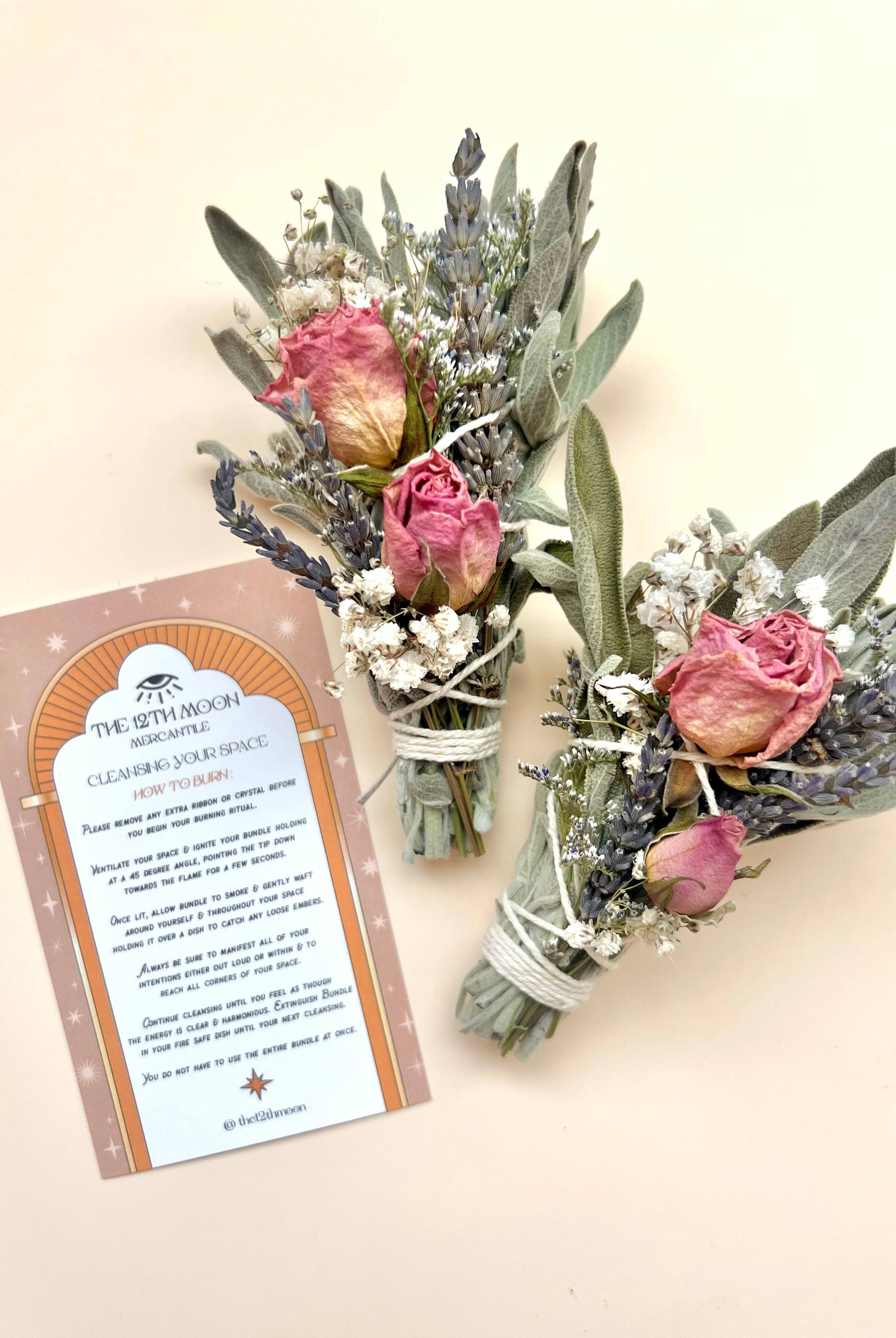 Two small floral bouquets with dried roses and lavender next to a MAGIC RITUALS Mini Sage Smudge Bundle - Smoke Cleanser on a pale background, infused with essential oil for aromatherapy benefits by The 12th Moon.