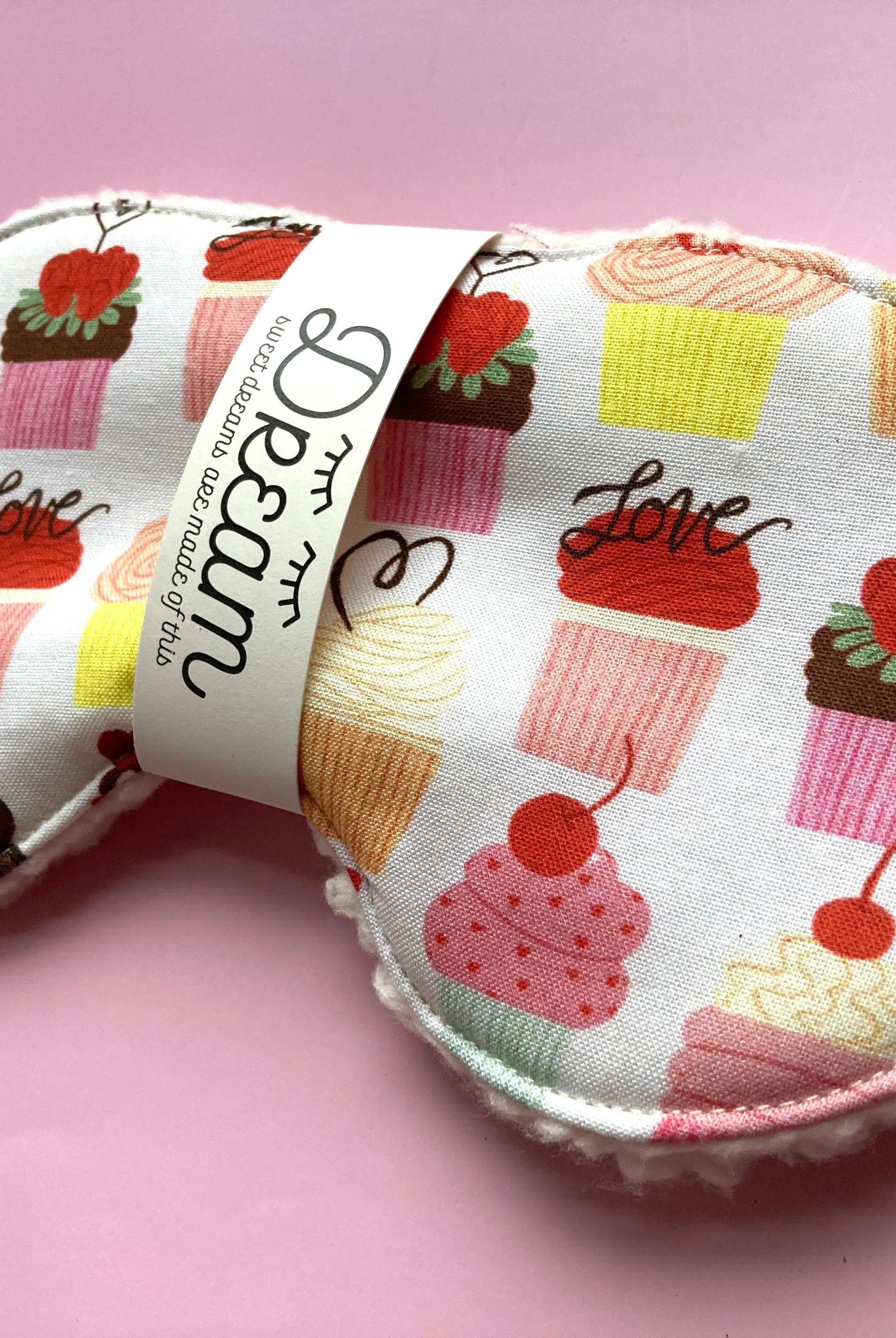 A Cupcake Sleep Mask by Little Man with a pattern of various cupcakes and the word "love" printed on it, infused with wild nurturer aromatherapy essential oil, displayed on a pink background.