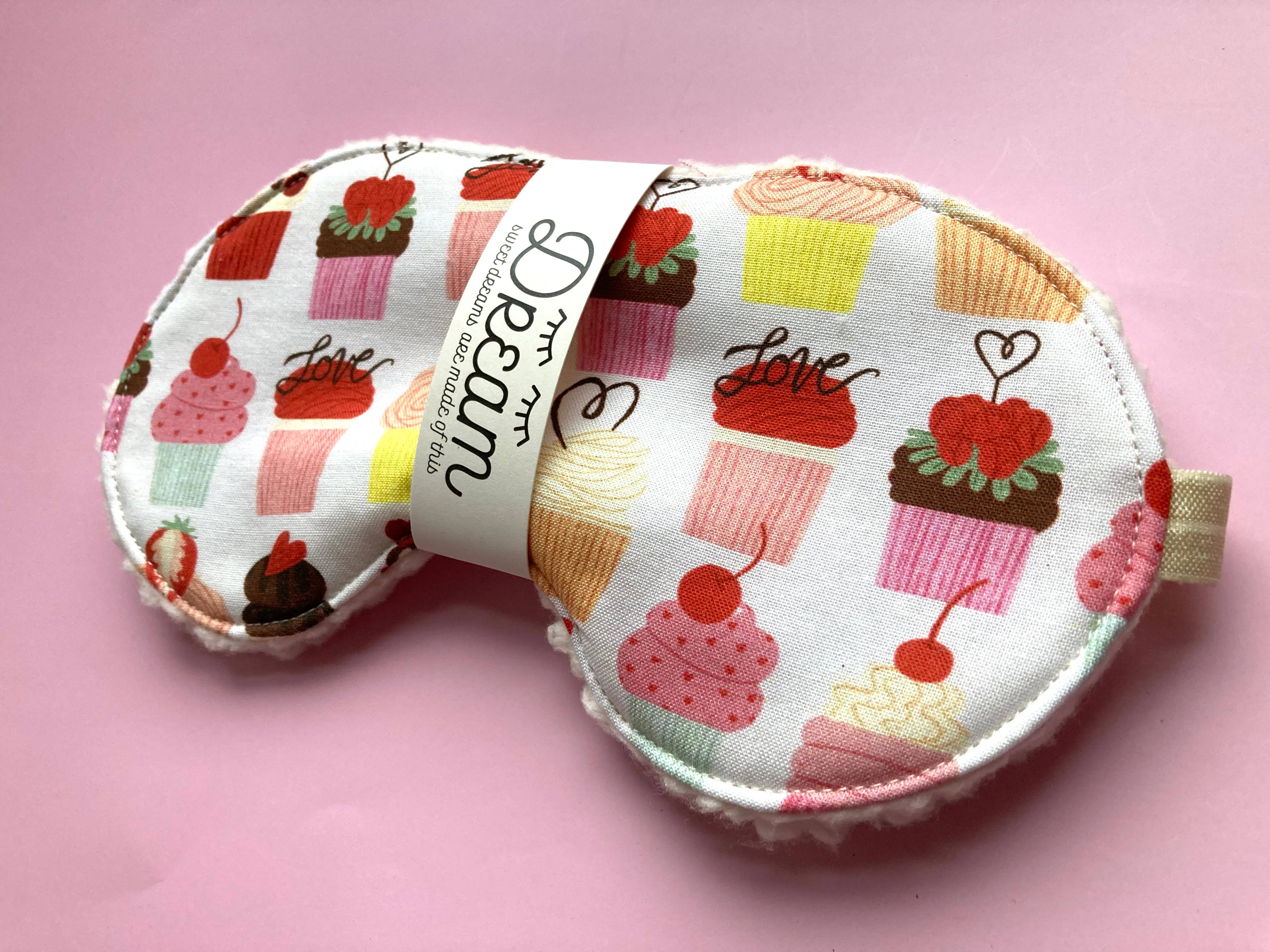 A Cupcake Sleep Mask by Little Man with a pattern of various cupcakes and the word "love" printed on it, infused with wild nurturer aromatherapy essential oil, displayed on a pink background.